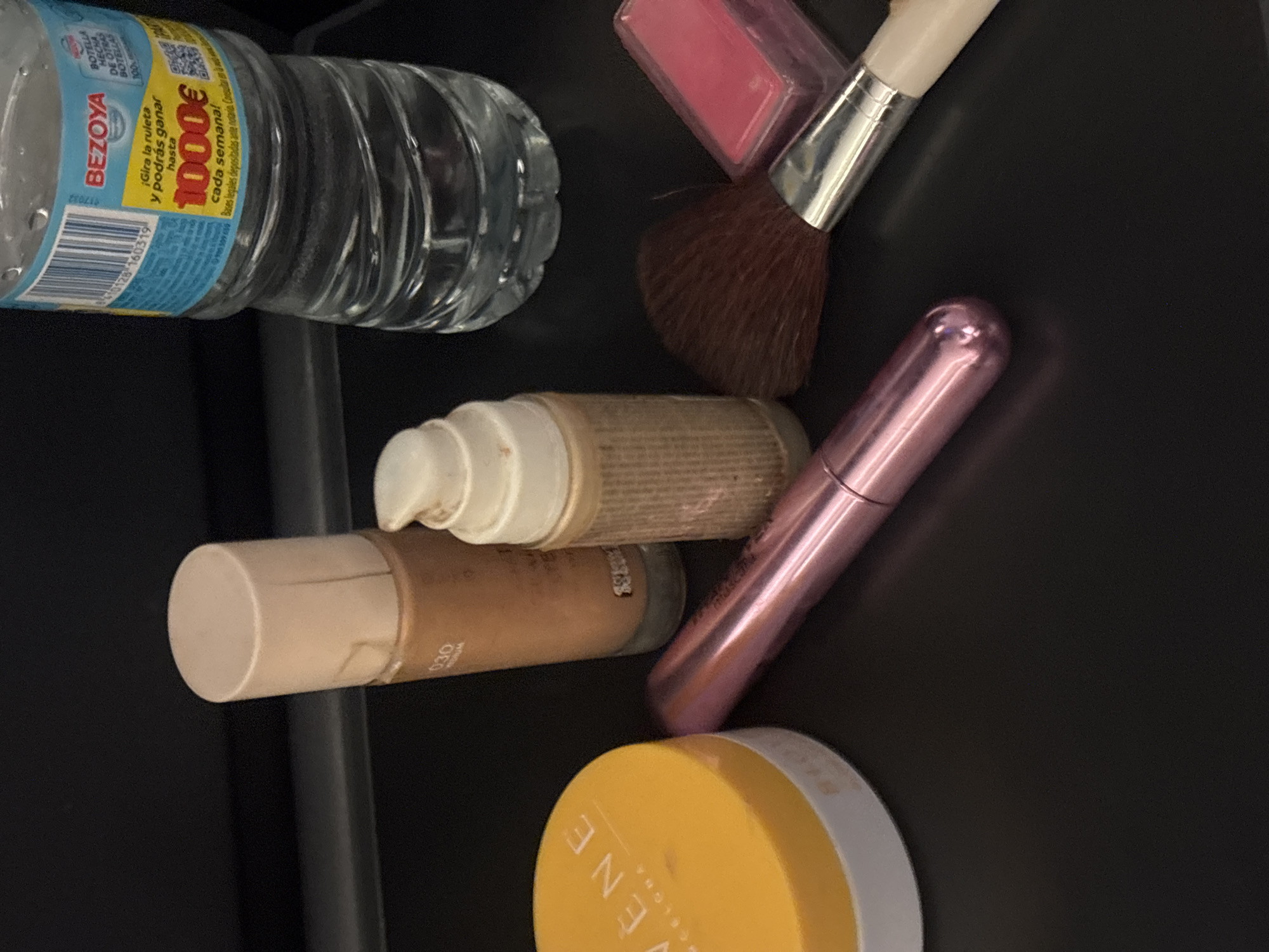 In Santa Maria de Palautordera, Spain, on a dark, flat surface, a collection of cosmetic items and a water bottle are arranged. From left to right, there is a yellow, round container with a white lid and faint text. Next to it, a tall, beige bottle with a beige cap and gold text. To its right, a smaller, textured beige bottle with a white pump dispenser. Further right, a pink metallic tube lies horizontally. To the right of the pink tube, a makeup brush with a dark brown bristled head and a silver ferrule with a white handle is positioned diagonally. Adjacent to the brush is a small, pink rectangular item, possibly blush. In the background, a partially visible clear plastic water bottle with a blue and yellow label is standing upright. The label on the bottle includes text in Spanish. The lighting suggests an indoor setting with diffused light. There are no people visible in the image.
