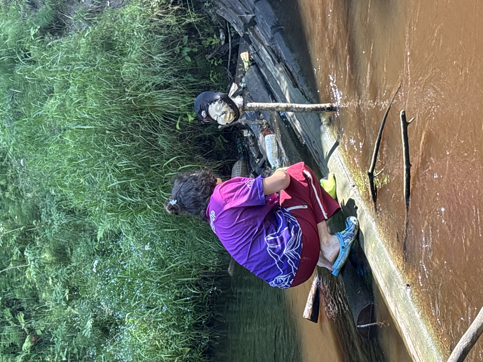 In Solnechnoye, Russia, on a sunlit day, a person kneels on a fallen log that spans a muddy, shallow river. The person, with curly dark hair pulled into a bun and wearing a purple t-shirt and red trousers, faces away from the camera, observing something in the water. Beside them, on a wooden structure that appears to be a makeshift bridge, a detached doll's head is impaled on a stick, its expression seemingly fixed in a grimace. The surrounding environment is lush and green, with tall grasses and foliage lining the riverbank. Debris, including plastic bottles and other refuse, is scattered on the wooden structure, suggesting a degree of neglect or disuse of the area.