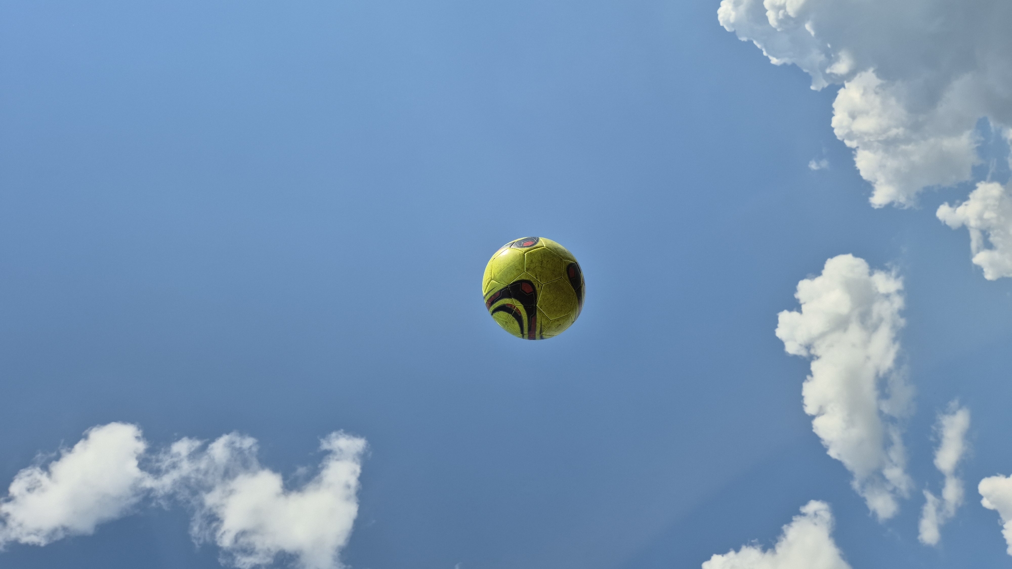 A soccer ball, predominantly yellow with black and red accents, floats in the clear blue sky. Scattered white clouds populate the upper and lower portions of the frame. The ball is positioned in the center of the image, slightly off-center to the left, and appears to be in mid-air.