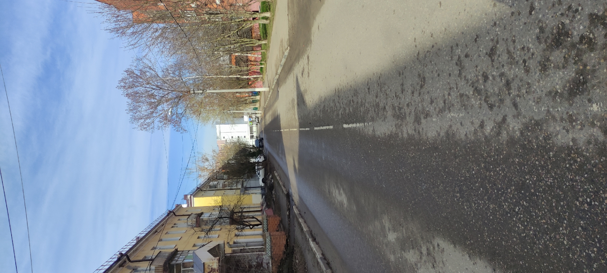 The image depicts a street in Fryazino, Russia. The sky is blue with sparse clouds. The street pavement is gray and damp, showing wear and tear. A broken white lane divider runs down the center of the road. On the left side of the street, there is a low-rise, light yellow residential building with visible windows and architectural details. Trees line both sides of the street. A power pole is visible on the right. In the distance, buildings can be seen along with parked cars.