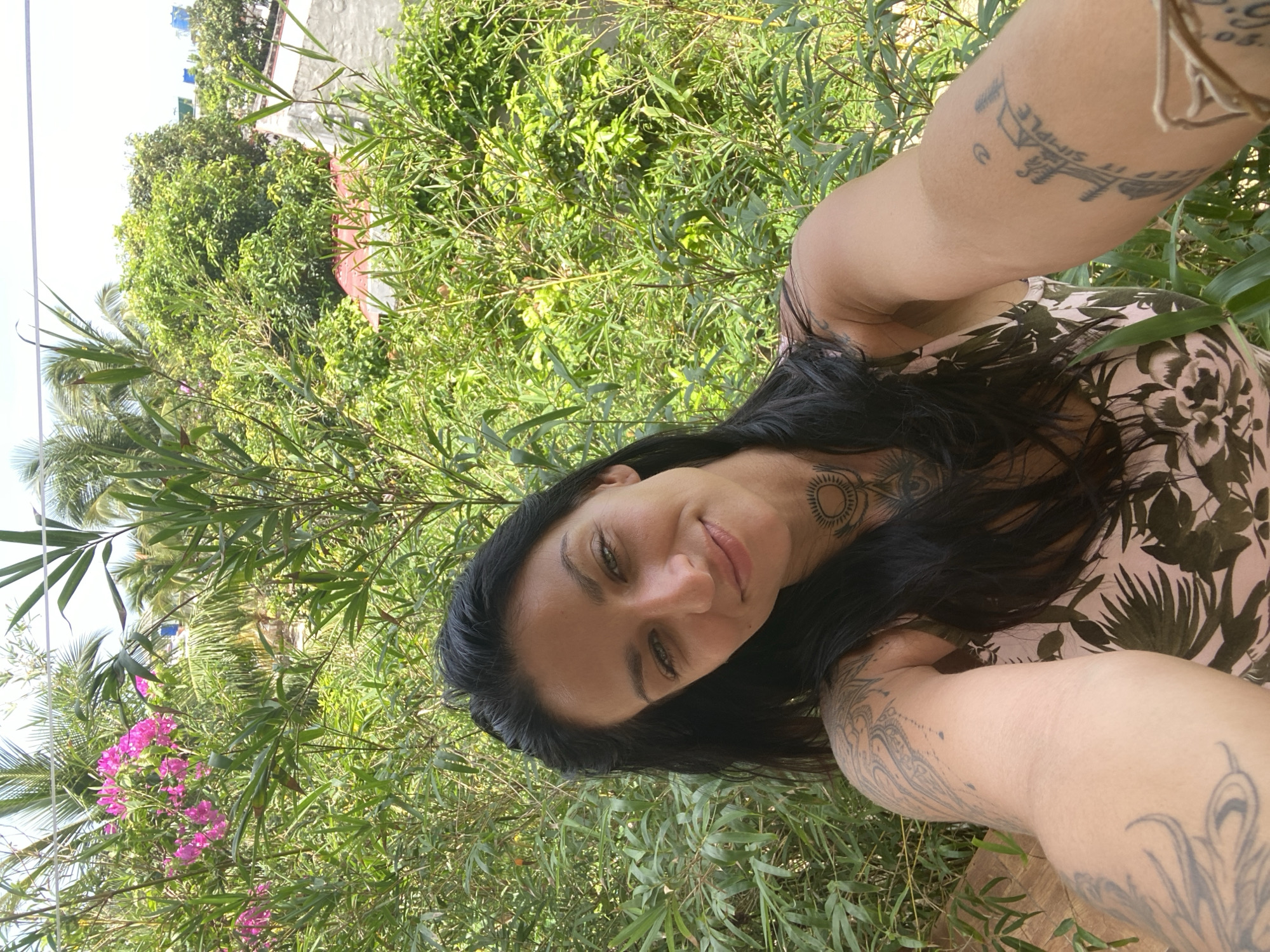 A woman with dark hair and green eyes smiles at the camera, holding it to take a selfie. She is outdoors, surrounded by lush green foliage. She has tattoos on her arms and neck. In the background, there are palm trees and a few buildings, suggesting a tropical location. The scene appears to be in Mũi Né, Vietnam, given the presence of tropical vegetation and architecture characteristic of the region.
