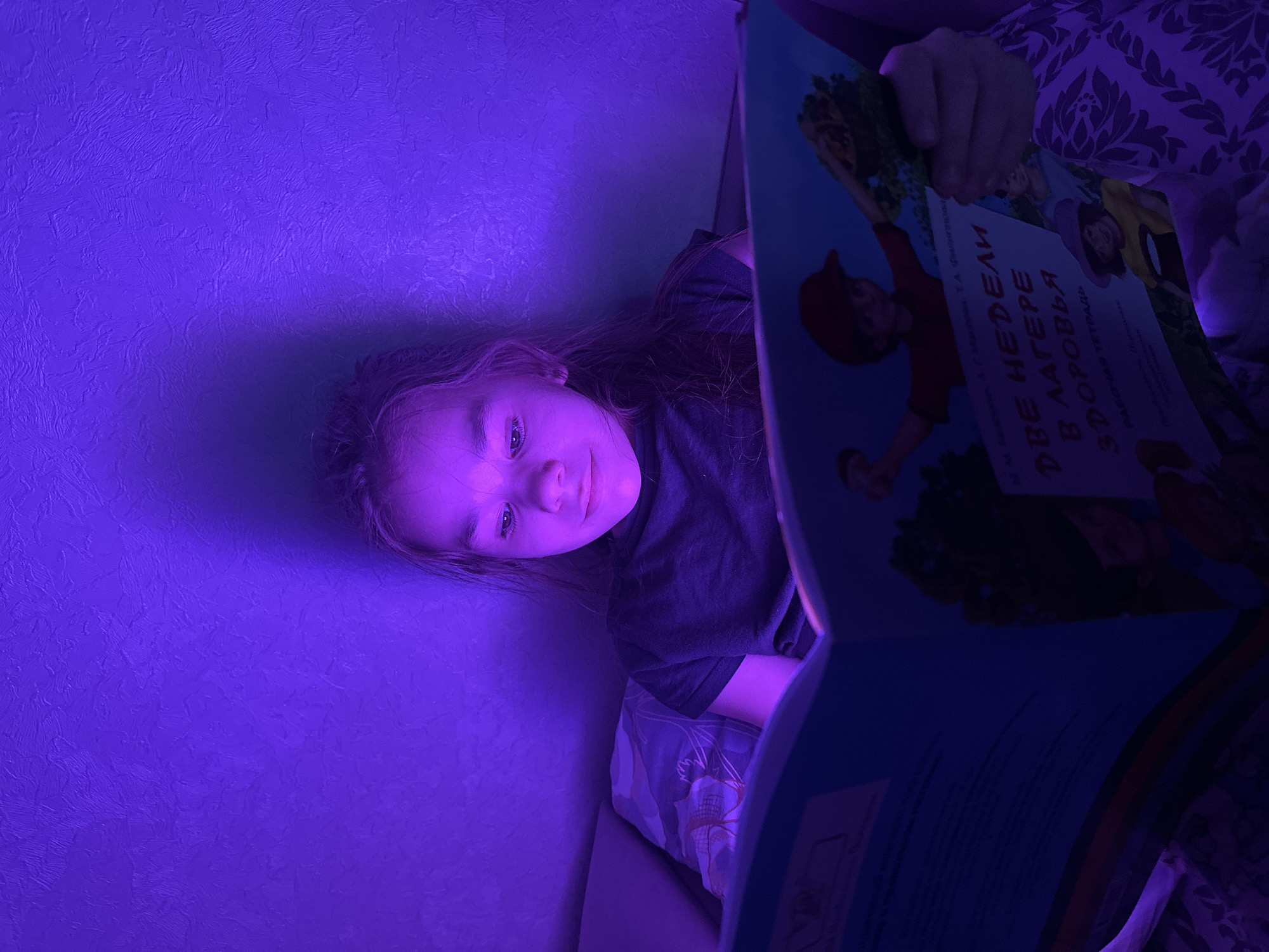 A young person, likely a girl with long dark hair, is depicted reading a book in a dimly lit room, illuminated by a strong purple light. The image is captured from a slightly rotated perspective, showing the girl lying on her side, looking towards the camera with a neutral expression. She wears a dark t-shirt, and a patterned fabric, possibly bedding, is visible beneath her.
The activity taking place is reading. The subject is holding an open book, partially obscuring the lower right portion of the frame. The wall behind her is a vibrant purple with a textured, stucco-like finish. The artificial, deep purple lighting suggests an evening or nighttime setting in Uva, Russia.
Visible text on the book cover is in Cyrillic: "ДВЕ НЕДЕЛИ В ЛАГЕРЕ ЗДОРОВЬЯ" which translates to "TWO WEEKS AT THE CAMP OF HEALTH." Below this, "РАБОЧАЯ ТЕТРАДЬ" meaning "WORKBOOK" or "ACTIVITY BOOK" is also visible, along with the author's name, "Т.А. Фисенко." The scene conveys a quiet, focused atmosphere.