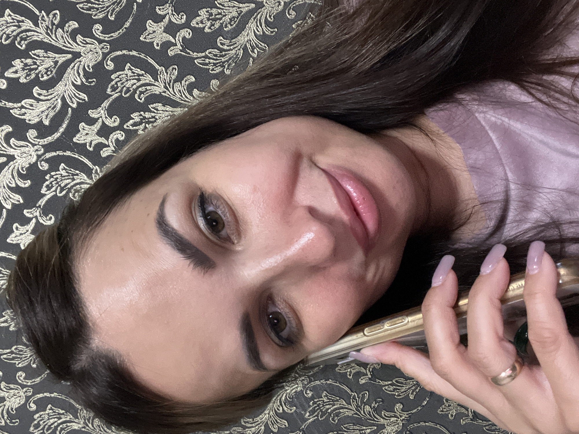 A woman with dark brown hair, fair skin, and brown eyes is holding a golden cell phone to her right ear and speaking. She is wearing a lavender shirt. Her fingernails are painted a light purple color, and she is wearing a gold ring on her left ring finger. The background is a dark wallpaper with a gold floral pattern. The lighting is soft and appears to be from an artificial source. The image is a close-up, head-and-shoulders shot.