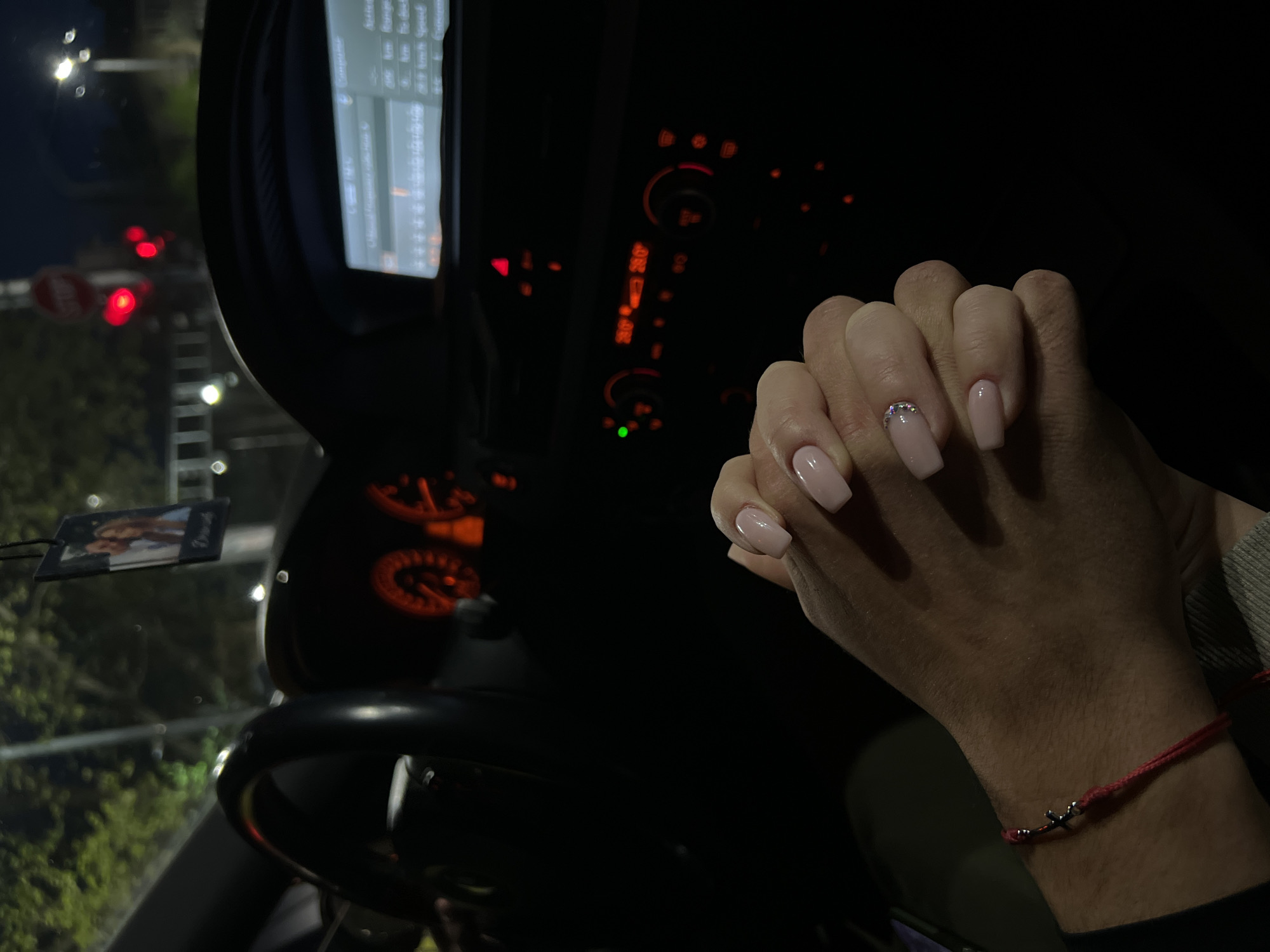 A photograph shows the interior of a vehicle, potentially a car, at night. The dashboard is visible with illuminated displays. The gear shift and steering wheel are also visible. The exterior view through the windshield shows lights, possibly from other vehicles or street lighting. The subject's hands are in the frame, wearing polished nails and a bracelet. A small hanging item dangles from the rearview mirror. The photograph was taken at night. The location is unknown, but the user suggests Plovdiv, Bulgaria.