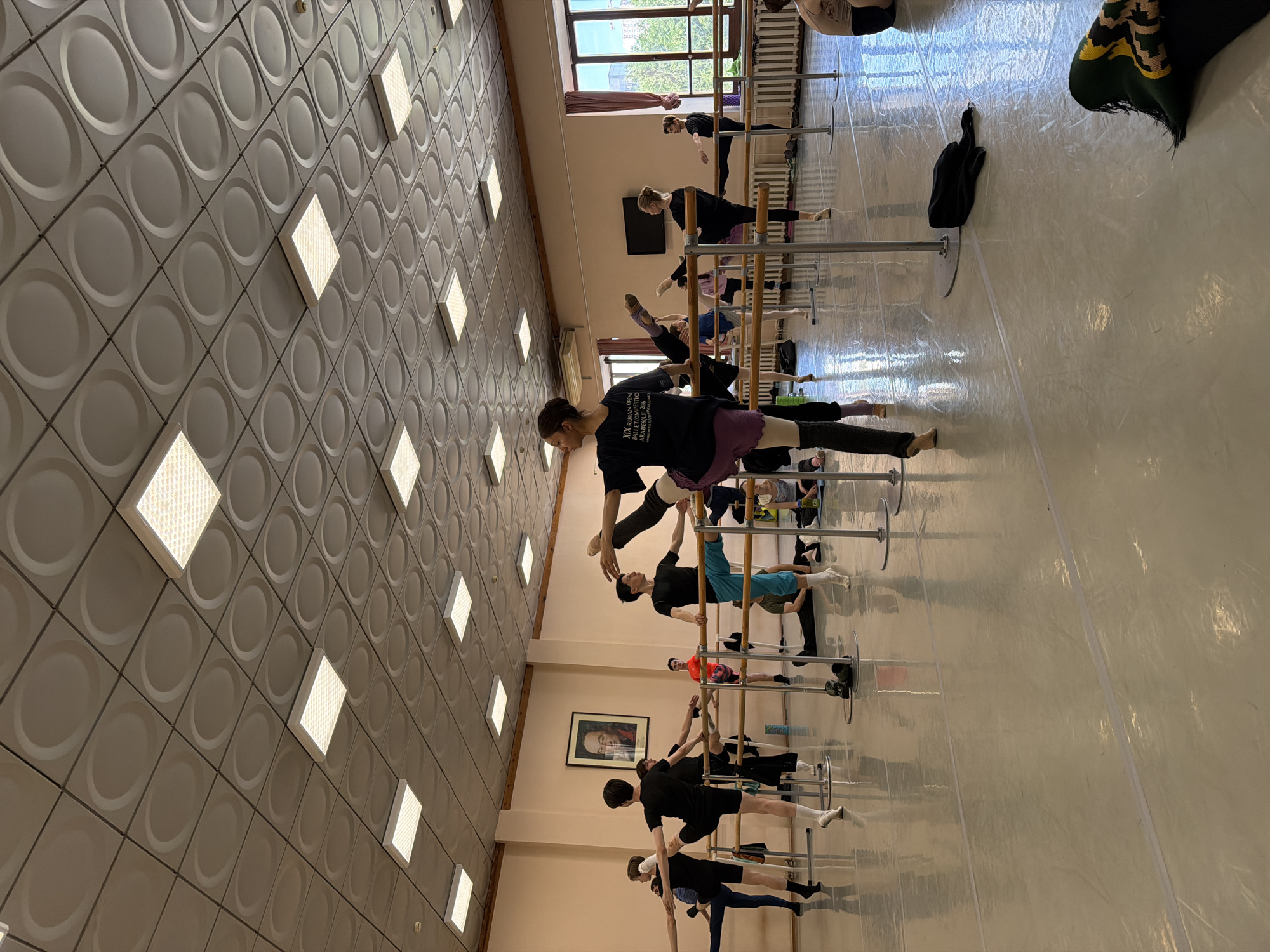 A ballet class is taking place in a studio, with several dancers practicing at ballet barres. The room features a distinctive ceiling with a grid of square panels, each containing a raised circular pattern, and illuminated by fluorescent lighting. A portrait of a man in a suit hangs on the pale pink wall. The floor is a reflective gray-white material. Dancers are dressed in leotards and tights, with some wearing t-shirts. The studio is equipped with multiple parallel ballet barres supported by metal poles. The scene captures a moment of focused physical activity within a dedicated training space.
