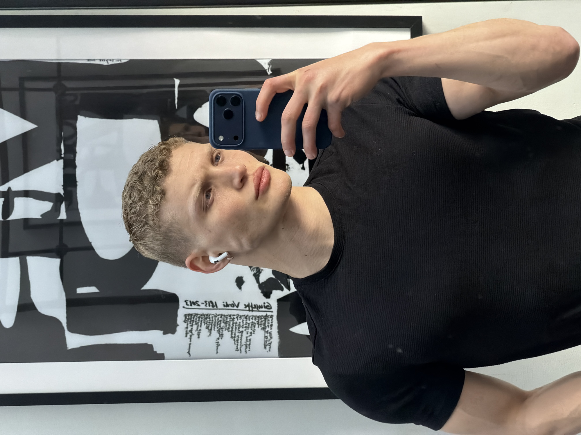 A young adult male with short, blond hair and light skin is holding a blue smartphone in his right hand, taking a selfie. He is wearing a black, ribbed short-sleeved shirt. An earpiece is visible in his left ear. Behind him is a black and white framed print on a white wall. The print appears to be an abstract design. The location is indoors, likely London, United Kingdom, suggested by the style and setting of the photograph.