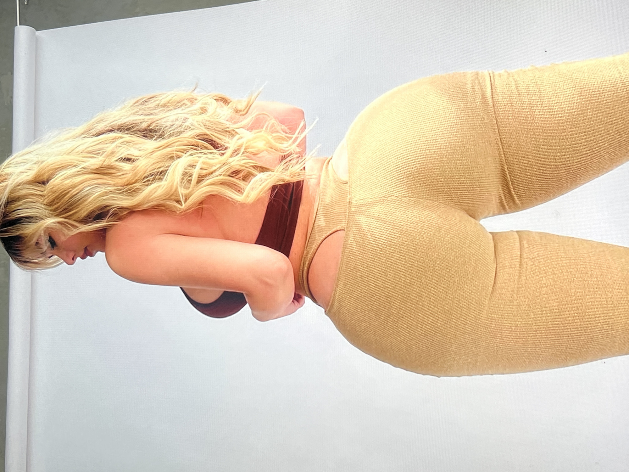 The media file shows a close-up, side-view of a blonde woman from the waist down. She is wearing gold, textured leggings with a cutout detail at the hip. Her hair is long and wavy, falling over her back. She is leaning forward, with her arms clasped in front of her. The background is a plain white studio backdrop, with a hint of a rolled white fabric on the left side, suggesting a professional photo shoot. The lighting appears to be bright and even, typical of a studio environment. No specific time of day, weather, or location cues beyond the studio setting are visible. The woman's expression is not fully visible, but her posture suggests a moment of repose or posing. There is no visible text in the image. The scene is static, with no discernible activity or event taking place other than the posed subject.