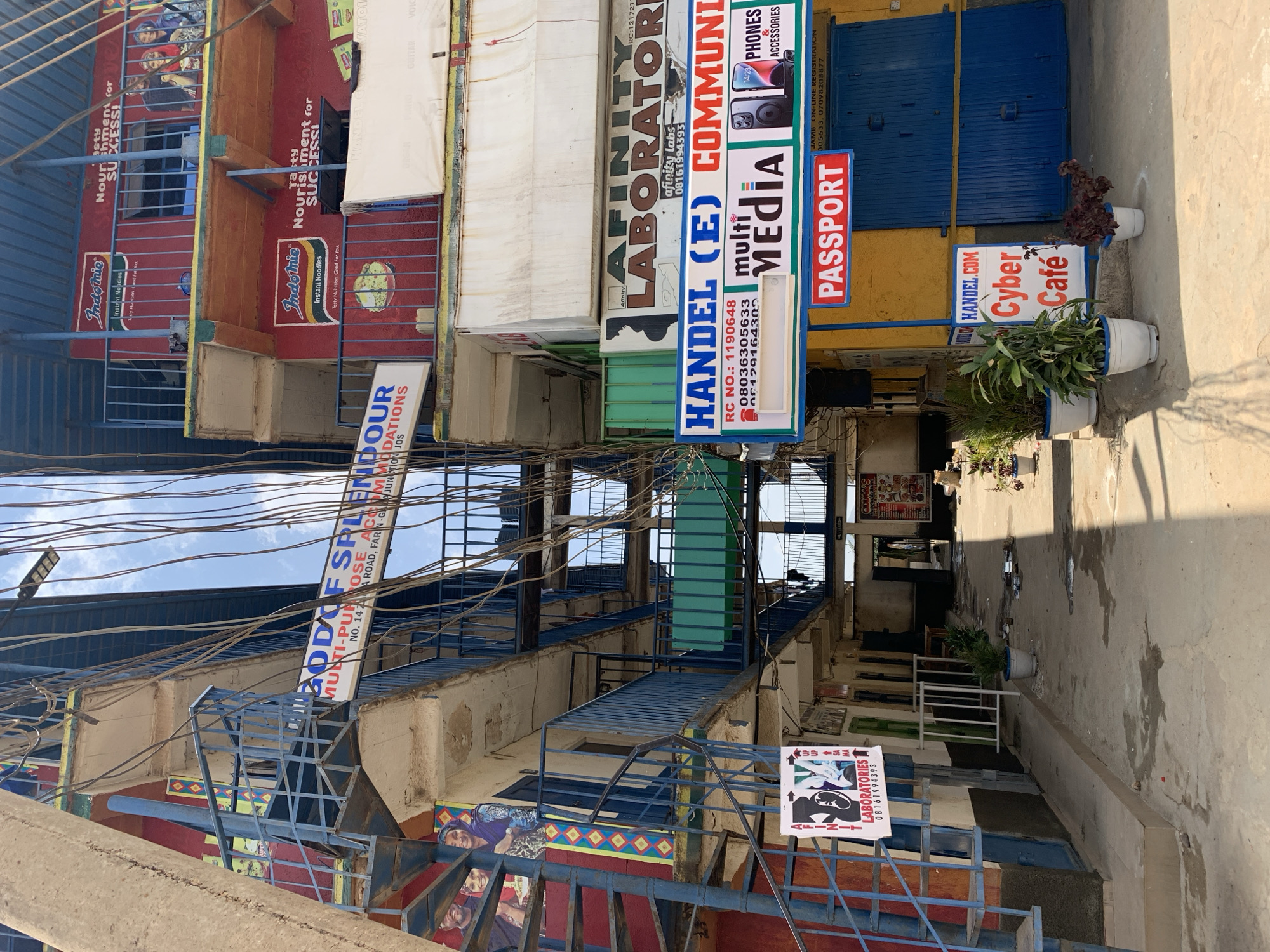 The image shows a cluttered urban street in Rafin Par, Nigeria. Multi-story buildings with exterior staircases line the narrow street. The buildings are covered in a variety of advertisements and signs, including for "Indomie" instant noodles, "Afinity Laboratories," "HANDEL (E) multi Media", a "Cyber Cafe", and "Passport". A sign also reads "God Of Splendour Multi-Purpose Accommodations". Overhead, a dense web of electrical wires obscures the sky. Several potted plants sit at the street level. The image does not show any people.