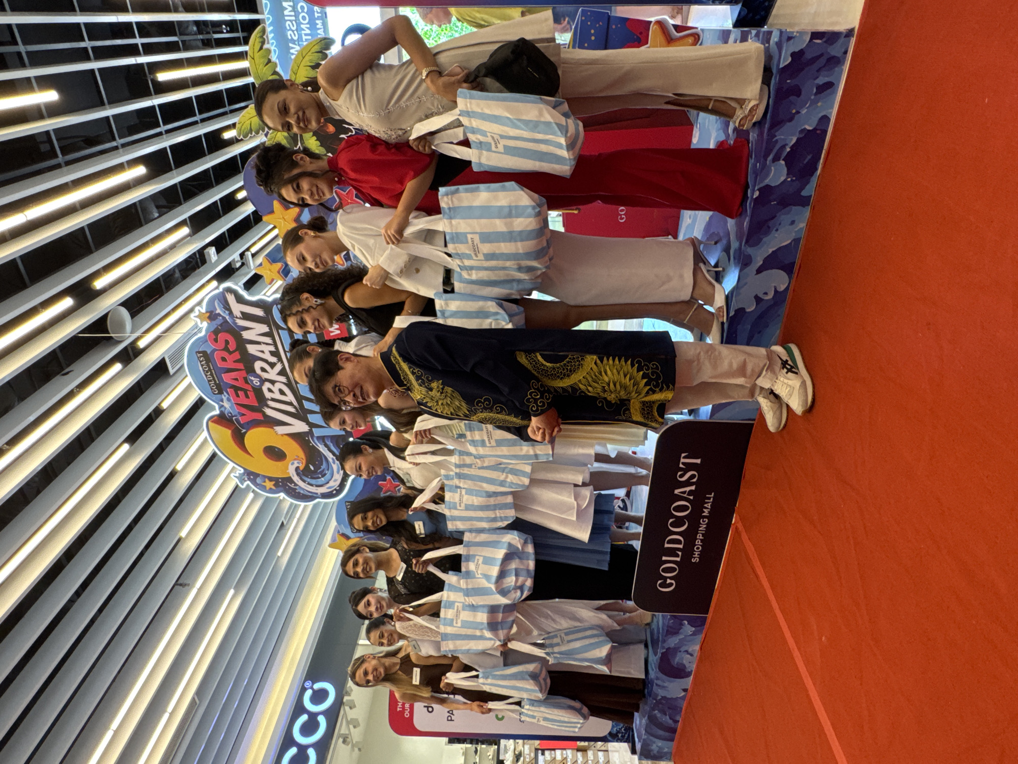 A group of ten women and one man are standing on a raised blue platform with a graphic of waves and stars. The women, dressed in formal attire and holding tote bags, appear to be models or contestants. The man, in traditional Vietnamese attire, is standing at the far right of the group. A banner behind them reads "GOLDCOAST 6 YEARS VIBRANT". The scene is set in an indoor shopping mall, indicated by the tiled ceiling and store signage visible in the background. A red carpet leads up to the platform. The event appears to be a promotional or celebratory gathering for the Goldcoast Shopping Mall. The location is specified as Nha Trang, Vietnam.