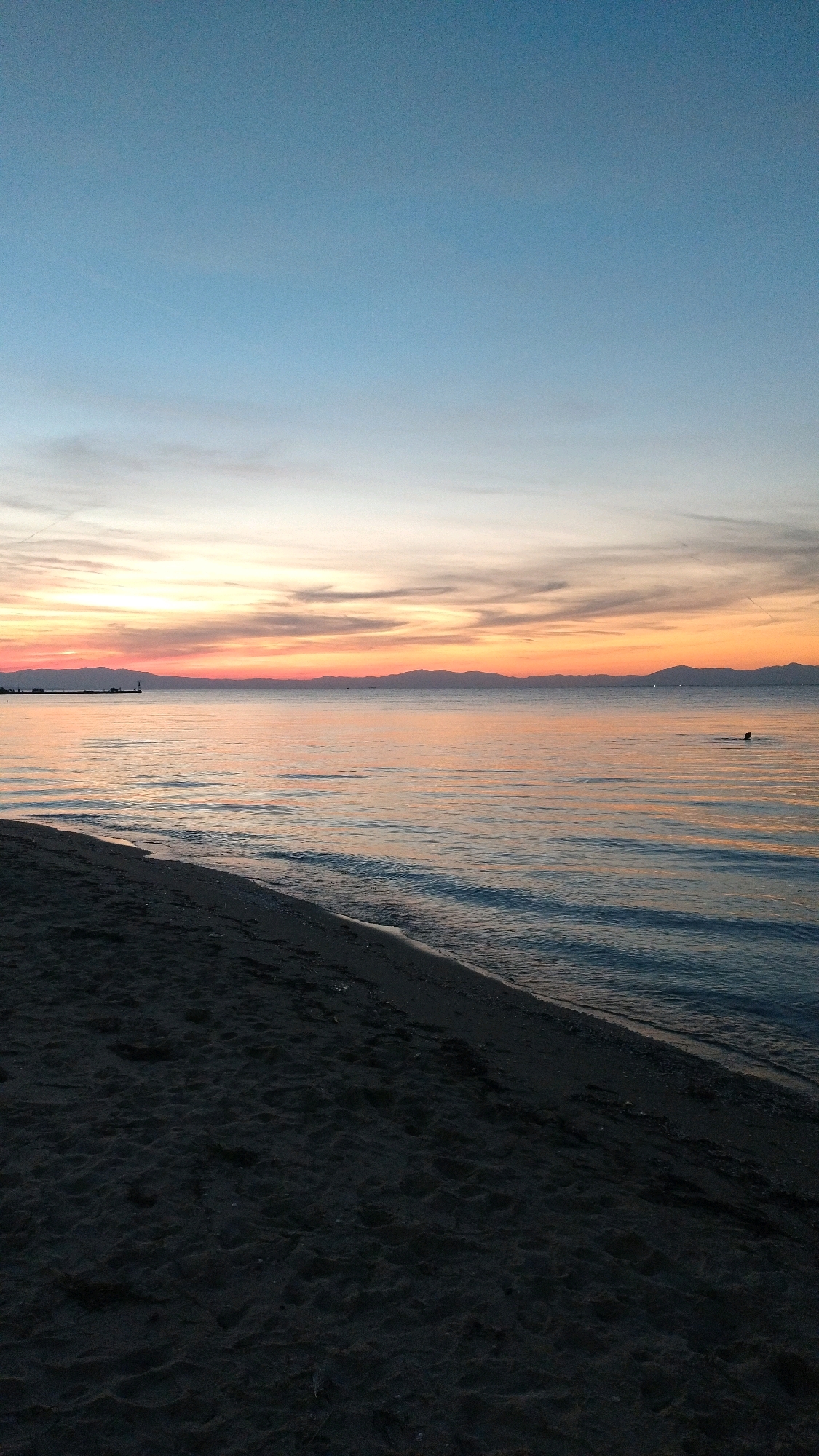 A photograph captures a serene sunset over the Thermaic Gulf from the beach in Perea, Greece. The sky transitions from a pale blue at the top to warm hues of orange, pink, and yellow near the horizon, streaked with wispy clouds. The calm sea reflects the colors of the sunset, creating a gentle shimmer on the water's surface. In the distance, a faint silhouette of mountains forms the coastline. A solitary figure is visible swimming in the sea on the right side of the image. The foreground is dominated by the dark, sandy texture of the beach, with subtle imprints and small pebbles visible. The edge of the water gently laps against the shore. A small lighthouse and a faint structure are visible on the left horizon.