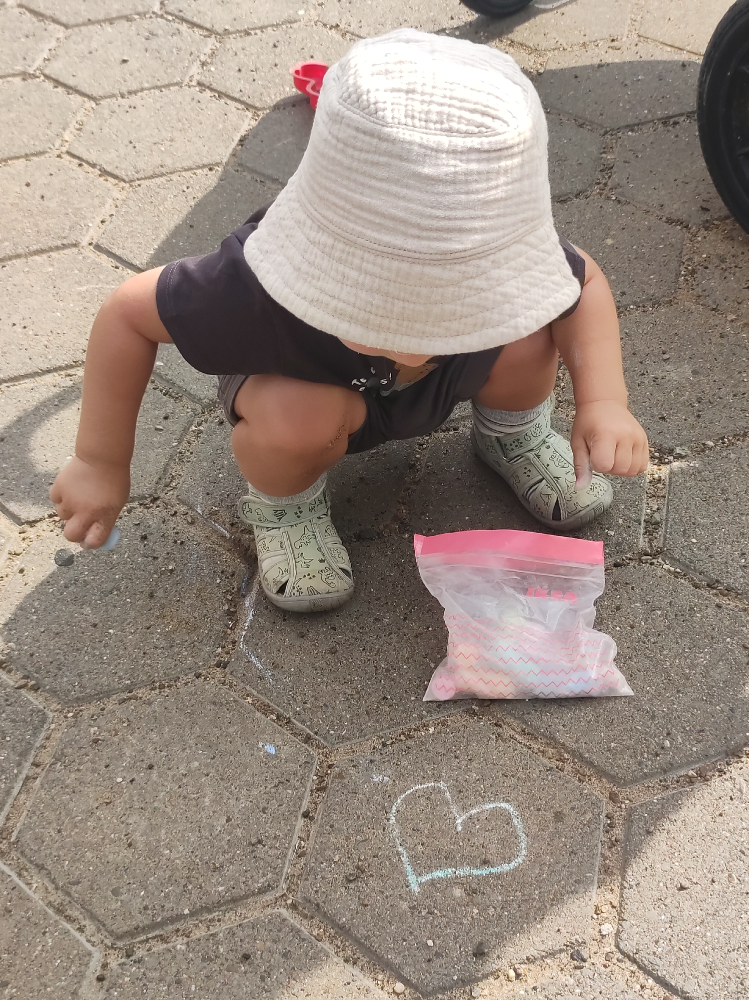 A young child wearing a white bucket hat, a dark t-shirt, grey shorts, grey socks, and light-colored sandals is crouching on a hexagonal paved surface. The child is holding a white object, possibly chalk, in their right hand and is drawing a heart shape with white chalk on the pavement. A clear plastic bag containing colorful chalk is located to the right of the child. The environment appears to be outdoors, with sunlight casting shadows on the ground. No specific location within Chaikovsky, Russia, can be determined from the image.