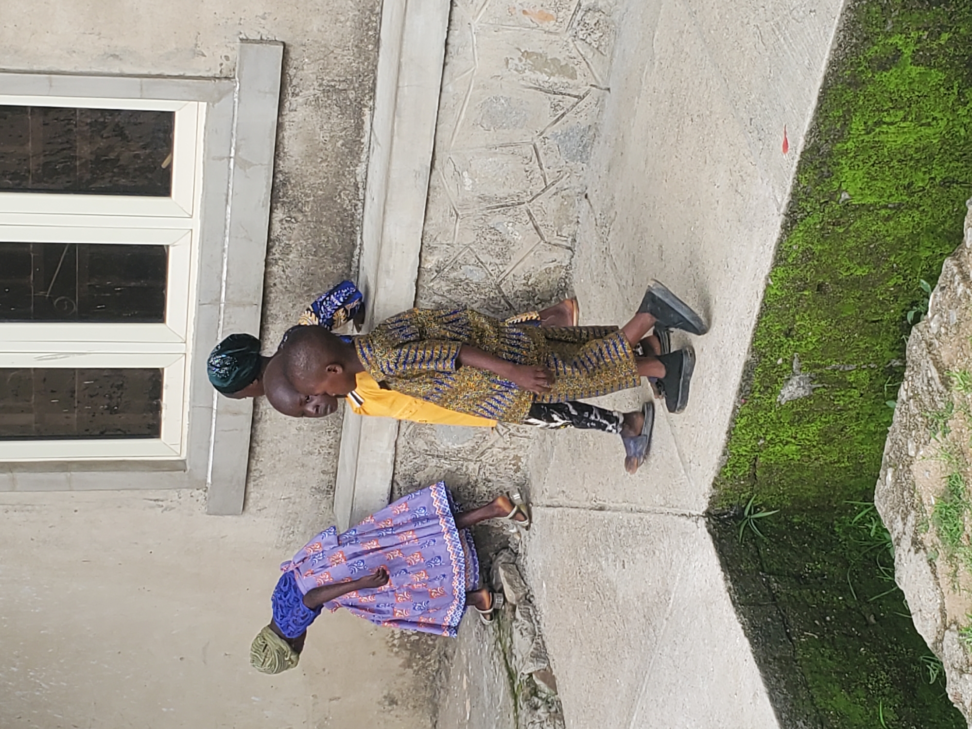 Three young children are walking along a concrete path in front of a building. The child on the left is wearing a purple dress with an orange and blue pattern and a green headscarf. They are bent over, with their head down. The middle child is wearing a yellow shirt and black and white patterned pants. The child on the right is wearing a yellow and blue patterned shirt and dark pants. They are all walking away from the viewer. The building behind them has a textured concrete wall and a white-trimmed window. To the right of the children, there is a moss-covered wall. The ground is a mix of concrete and some stones.