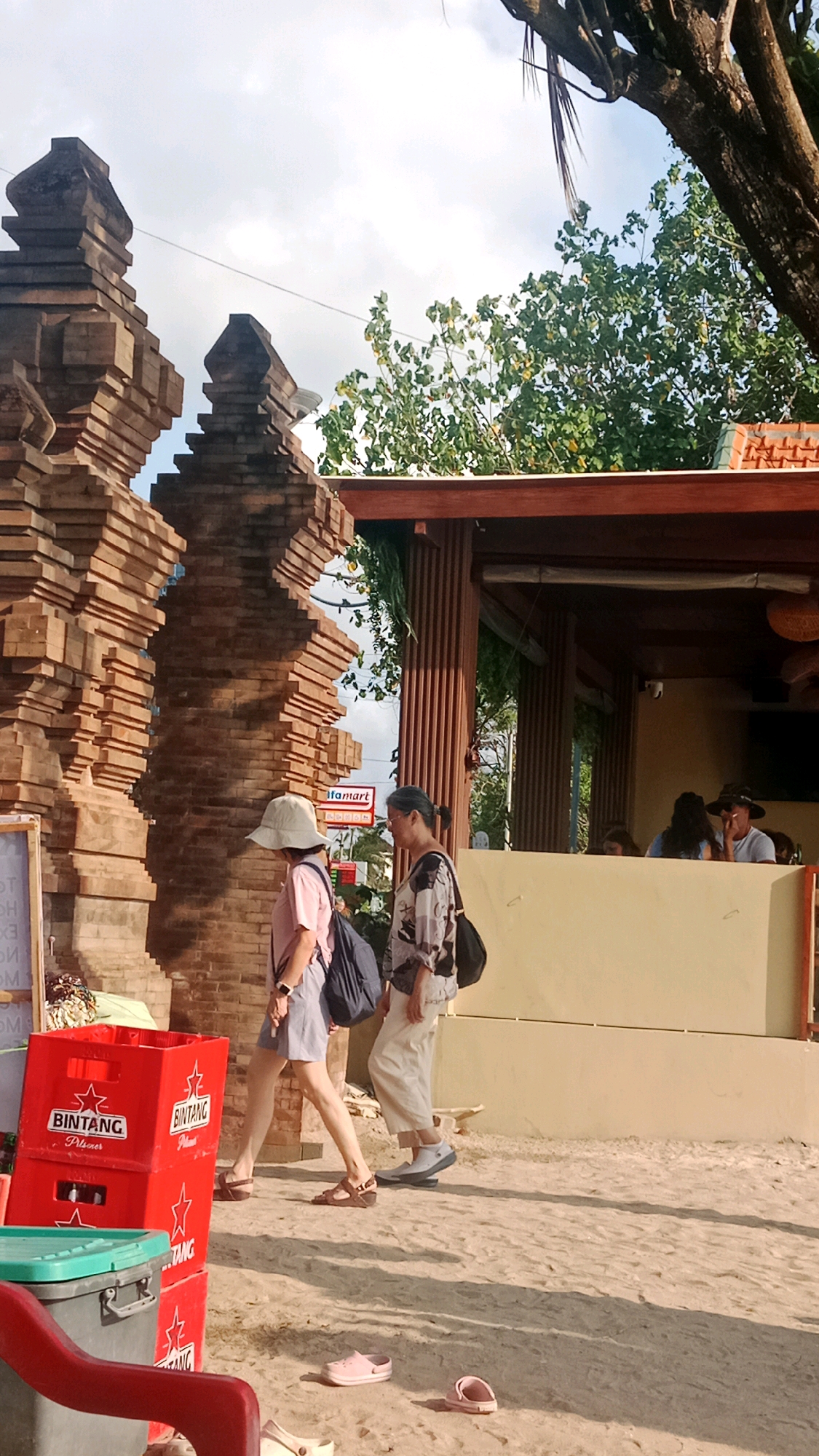 Two women are walking on a sandy path in front of a building with an open-air cafe area. The woman on the left wears a light-colored hat, a pink t-shirt, and gray shorts. She carries a dark blue backpack and brown sandals. The woman on the right wears a patterned shirt and cream-colored pants, with a black bag slung over her shoulder and white shoes. In the background, there is a brick structure resembling a gateway. Red crates labeled "Bintang" are stacked to the left of the path. The building has a wooden overhang and wooden vertical supports. Other people are visible inside the cafe. The ground is covered in sand and shadows.
