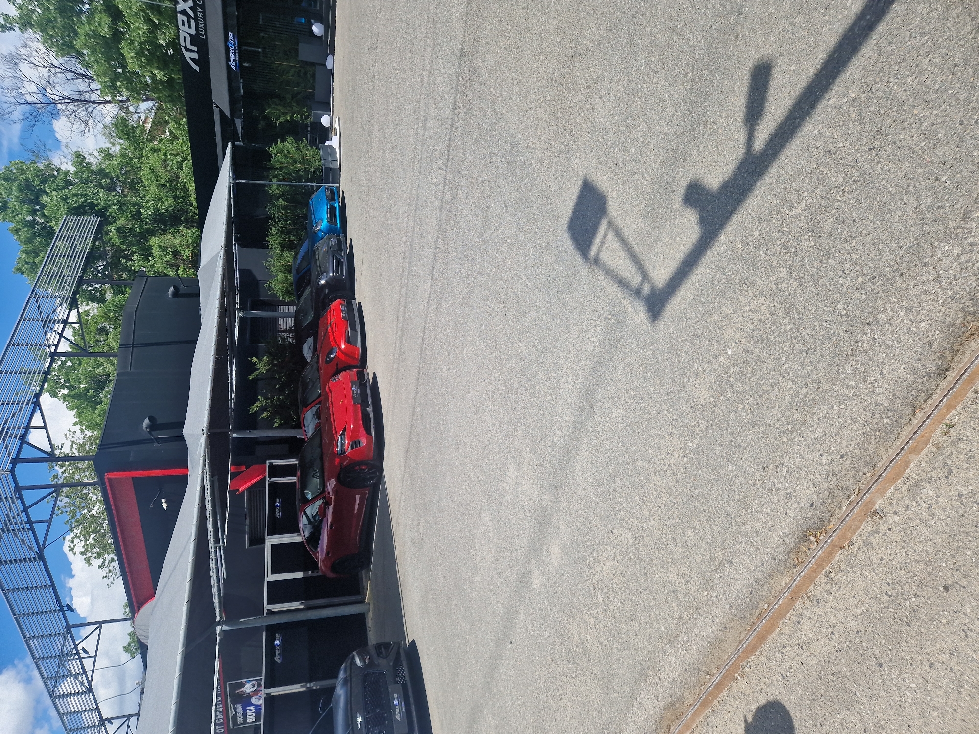 The image shows a parking lot, with a row of parked cars near a building. The cars are mostly red, with a few black and blue ones also visible. The building has the words "Apex Luxury Cars" on it. The sky is visible in the upper left corner, with some trees. The shadows of a light post is visible on the parking lot pavement. The location in the city of Unknown, Unknown is not discernible in the provided image.
