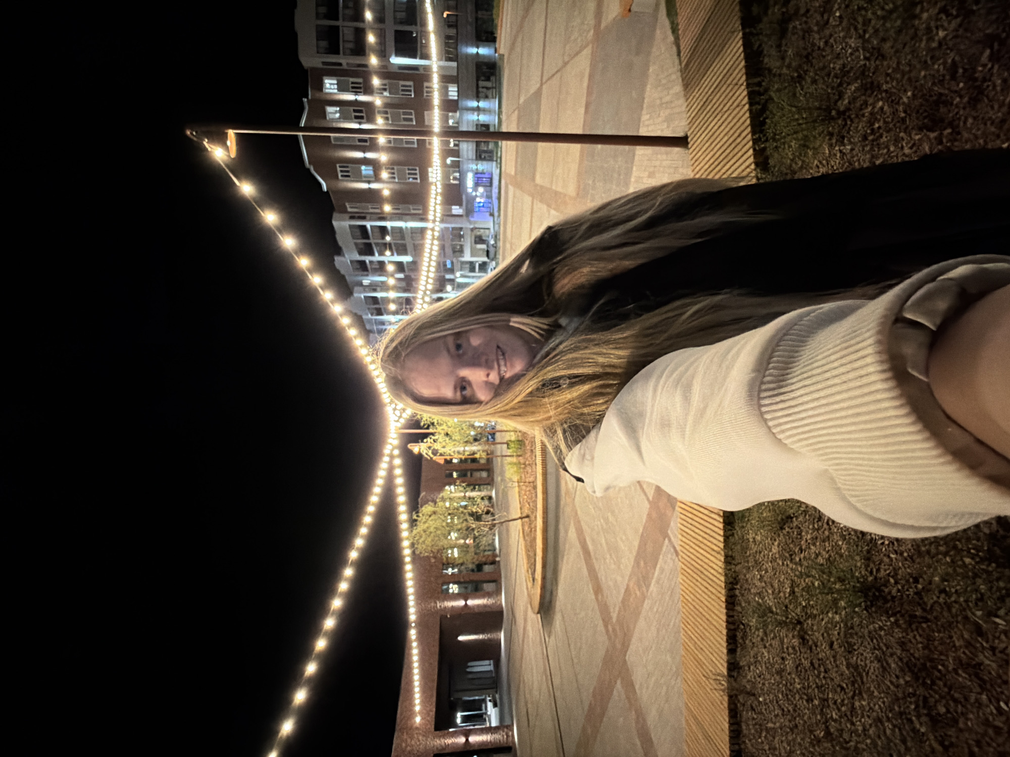 A young woman takes a selfie at night in Izhevsk, Russia. She is seated on a wooden bench, a building with lit windows is in the background, and string lights hang overhead. The ground is paved and bordered by grass. The woman wears a white sweater and dark outerwear.