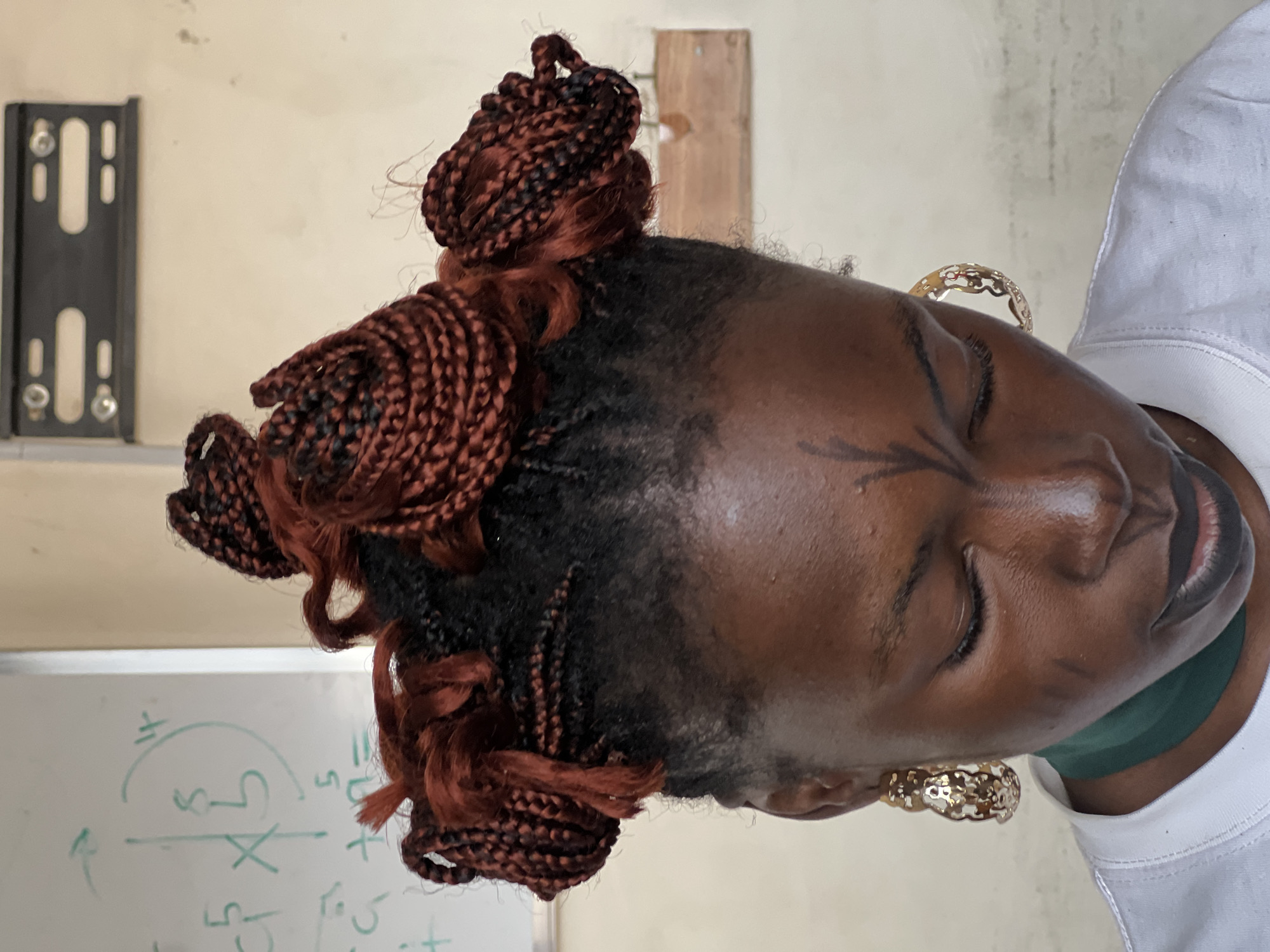A close-up, head-and-shoulders shot features a young Black woman from the chest up, her face turned downwards and to the right. Her dark skin is smooth, with some visible markings on her forehead and cheeks. A thin, dark green band is worn around her neck, and she has large, ornate gold hoop earrings. Her lips are painted a dark color, and her eyes are closed. Her hair is styled in numerous dark red braids, some of which are woven into four large, coiled buns atop her head. The background is a plain, light-colored wall, with a whiteboard on the left displaying green markings that appear to be mathematical equations. A black bracket is mounted on the wall above and to the right of her head. The scene appears to be indoors, possibly in a classroom or office setting, in Jos, Nigeria.