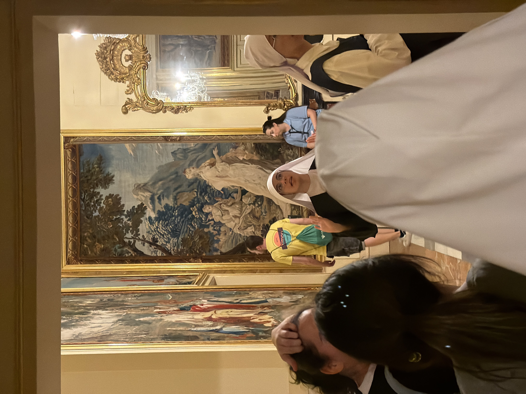 In the Hall of the Four Rooms within the Royal Site of San Lorenzo de El Escorial, Spain, a group of people are gathered, appearing to be tourists. The room is adorned with large, ornate tapestries depicting classical scenes. In the foreground, two women wearing nun's habits are visible, one in focus and another slightly out of focus. To the left, a person's hand is seen touching their head. Further back, a man in a yellow t-shirt with a green backpack walks away from the camera. Another woman, dressed in a blue shirt, is seated in the midground, looking towards the right. The environment suggests an indoor museum or historical building, with gilded frames on the tapestries and ornate architectural details visible in the background, including a large, decorative mirror. The lighting appears to be artificial, casting a warm glow on the scene.