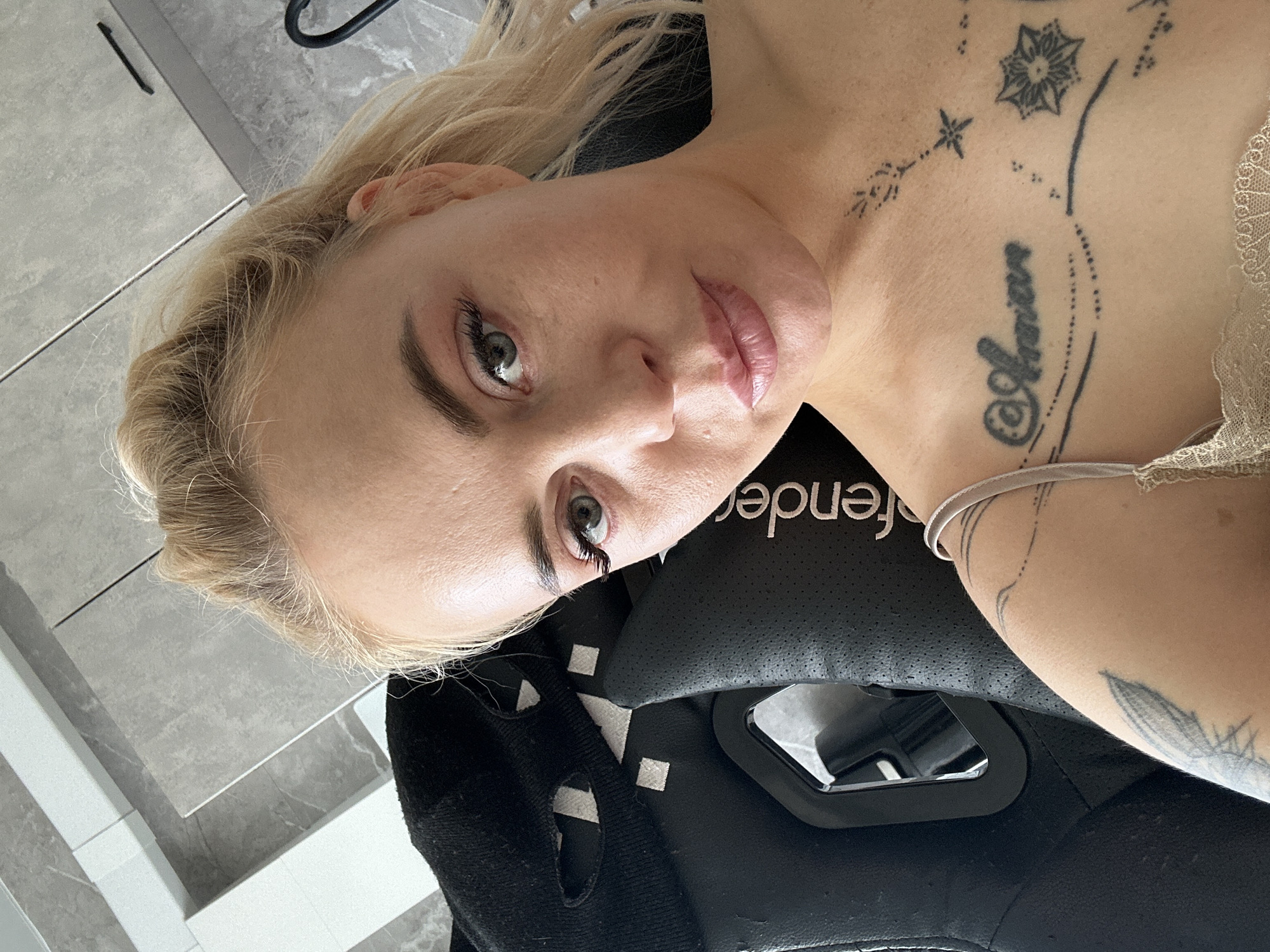 The image is a head-and-shoulders selfie of an adult female in an indoor, residential setting, specific location Unknown, Unknown. The subject, with blonde hair tied back and light green-blue eyes, looks directly at the camera with a neutral expression. She has fair skin, defined eyebrows, and full lips with a light pink gloss. She wears a light-colored, lace-trimmed camisole or bralette, with a thin strap visible on her right shoulder. Prominent black tattoos are visible on her upper chest, including text resembling "@Anvian" accompanied by dotted lines and star-like motifs. A partial tattoo resembling a feather or leaf is on her upper left arm. She is seated in a black, perforated faux-leather ergonomic or gaming chair, partially revealing the brand name "Defender" on the right side. The background features modern grey-toned cabinets or wall panels and what appears to be grey tiled flooring with white trim, indicating a contemporary interior design. A black hanging object is partially visible on the far right.