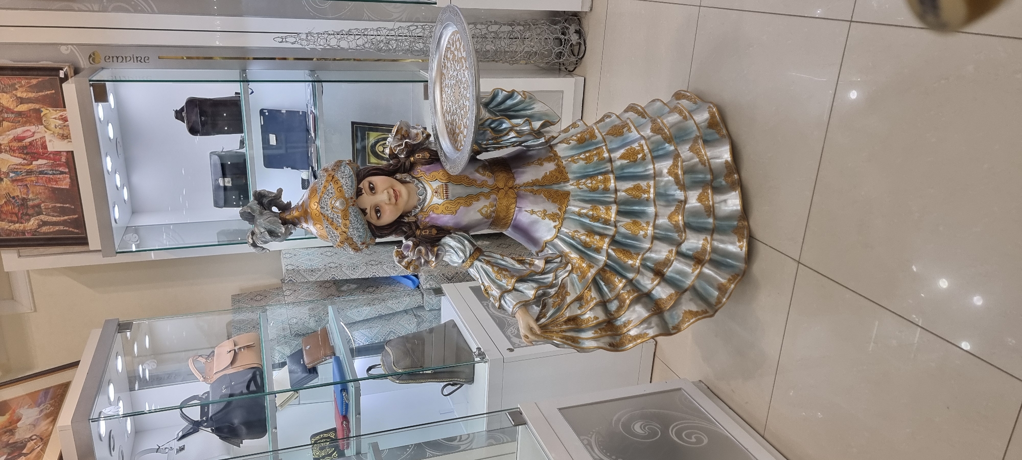 A full-body statue of a young girl in traditional attire stands in the center of a retail store, likely a boutique selling accessories or gifts. The statue depicts a girl with dark hair, a fair complexion, and dark eyes, wearing an ornate, multi-layered dress in shades of purple, silver, and gold. Her dress features intricate gold embroidery and ruffles, and she is adorned with a matching headdress. The girl is holding a decorative silver tray with a circular pattern in her right hand.
The background of the image reveals several glass display cases filled with various items, including handbags, wallets, and what appears to be a backpack. The display cases are lit from above, illuminating the products within. The overall environment suggests a well-organized and brightly lit retail space. The floor is tiled with large, light-colored square tiles. The signage visible on the display case on the right side of the statue reads "empire" vertically, suggesting the store's name. The presence of traditional attire on the statue, along with the store's name, could indicate a shop specializing in ethnic or cultural goods. While the specific location in Aktobe, Kazakhstan, is not explicitly stated in the image, such a store would be consistent with retail establishments found in urban areas of Kazakhstan.