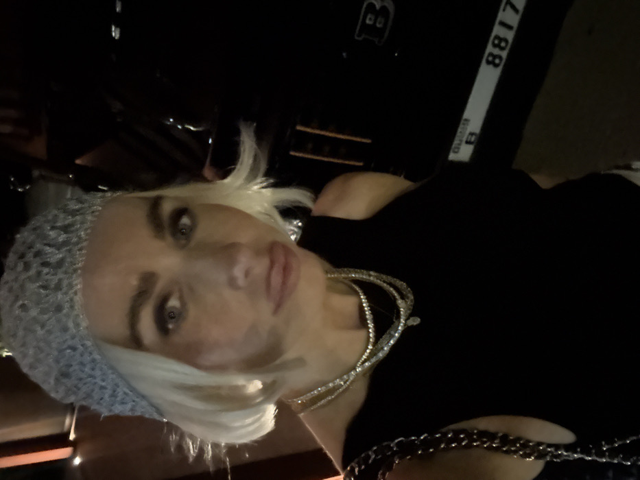 A fair-skinned woman with short blonde hair, wearing a grey textured hat and a black sleeveless top, is centered in a dimly lit, close-up self-portrait. She has light-colored eyes, prominent eyebrows, and her lips are slightly pursed in a pout. Multiple layered necklaces, appearing to be beaded and possibly pearl-like, adorn her neck, and a dark shoulder bag with a metal chain strap is visible on her left side. To the right of the woman, the front portion of a black vehicle is visible, featuring a distinct "B" logo, identifying it as a Brabus model. Below the logo, a Dubai license plate is discernible, displaying "Dubai" in English text along with the plate designation "B 8817". The scene is set at night in the city of Dubai, United Arab Emirates, with a warm light source visible on the far left of the frame.