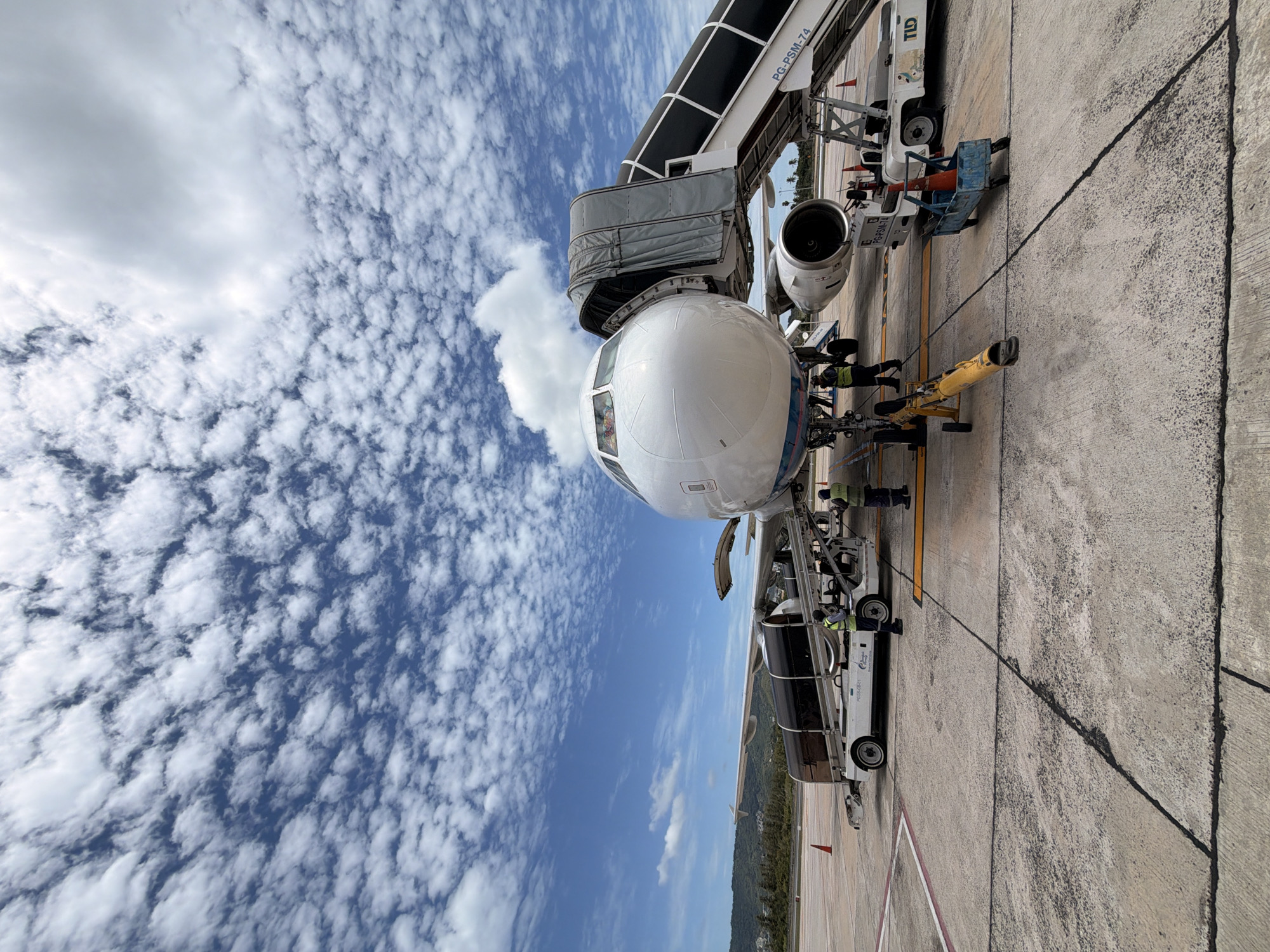 A wide-angle, low-angle shot captures an aircraft parked on a tarmac under a dramatic, cloud-filled sky in Ko Samui District, Thailand. The front of a white passenger airplane dominates the frame, with its nose cone and cockpit visible. A gray airstair is attached to the plane's fuselage, extending towards the ground. Several airport service vehicles, including a baggage loader and a small cart with cones, are positioned around the aircraft. Two individuals in high-visibility vests are present near the baggage loader, appearing to be working. The tarmac is marked with lines and has visible cracks and patches of wear. In the background, a lush, green hillside with trees and a distant coastline can be seen. The sky is filled with numerous small, puffy white clouds scattered across a bright blue expanse.