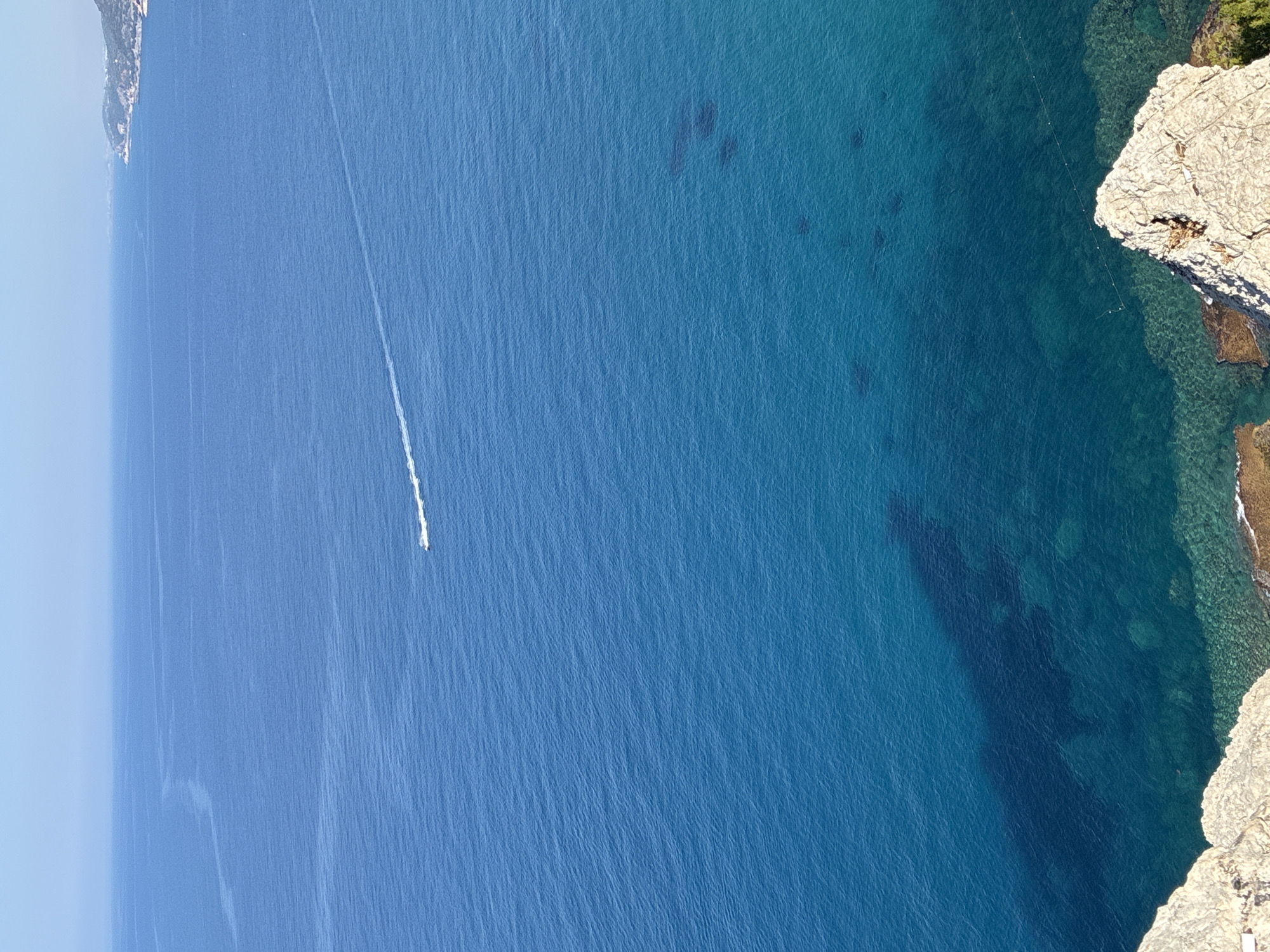 An aerial shot captures a vast expanse of the deep blue Mediterranean Sea under a clear sky. The water exhibits varying shades of blue, transitioning from a lighter hue near the horizon to a deeper, more translucent turquoise closer to the shore, revealing the seabed in some areas. A white boat is seen in the distance, leaving a foamy wake as it traverses the water. To the far right, a coastline dotted with vegetation and buildings is visible, suggesting a populated area. The foreground is dominated by the rugged, rocky surface of a cliff. The overall scene is serene and highlights the natural beauty of the coastal landscape. The image is consistent with the coastal region of Alanya, Turkey, known for its stunning Mediterranean views and cliffside overlooks.