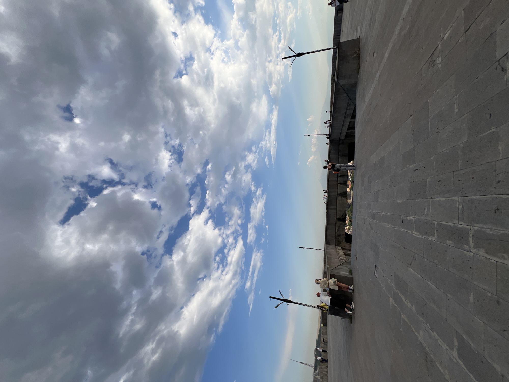 The image depicts a broad, paved plaza with a cloudy sky overhead. In the middle ground, several people are seen standing and walking along a concrete barrier that overlooks a lower level or sunken area, possibly an entrance to a structure or a viewpoint. Two individuals, a man and a woman, are positioned on the left side of the barrier, leaning against it and looking outwards. Another person is walking away from the camera on the right side of the barrier. In the distance, a tall, slender tower with a triangular top is visible on the left, and a distant cityscape with mountains can be seen on the horizon. The plaza is constructed from large, rectangular stone slabs, and a few streetlights or poles are present. The overall scene suggests a public space in Yerevan, Armenia, likely a viewpoint or a public square.