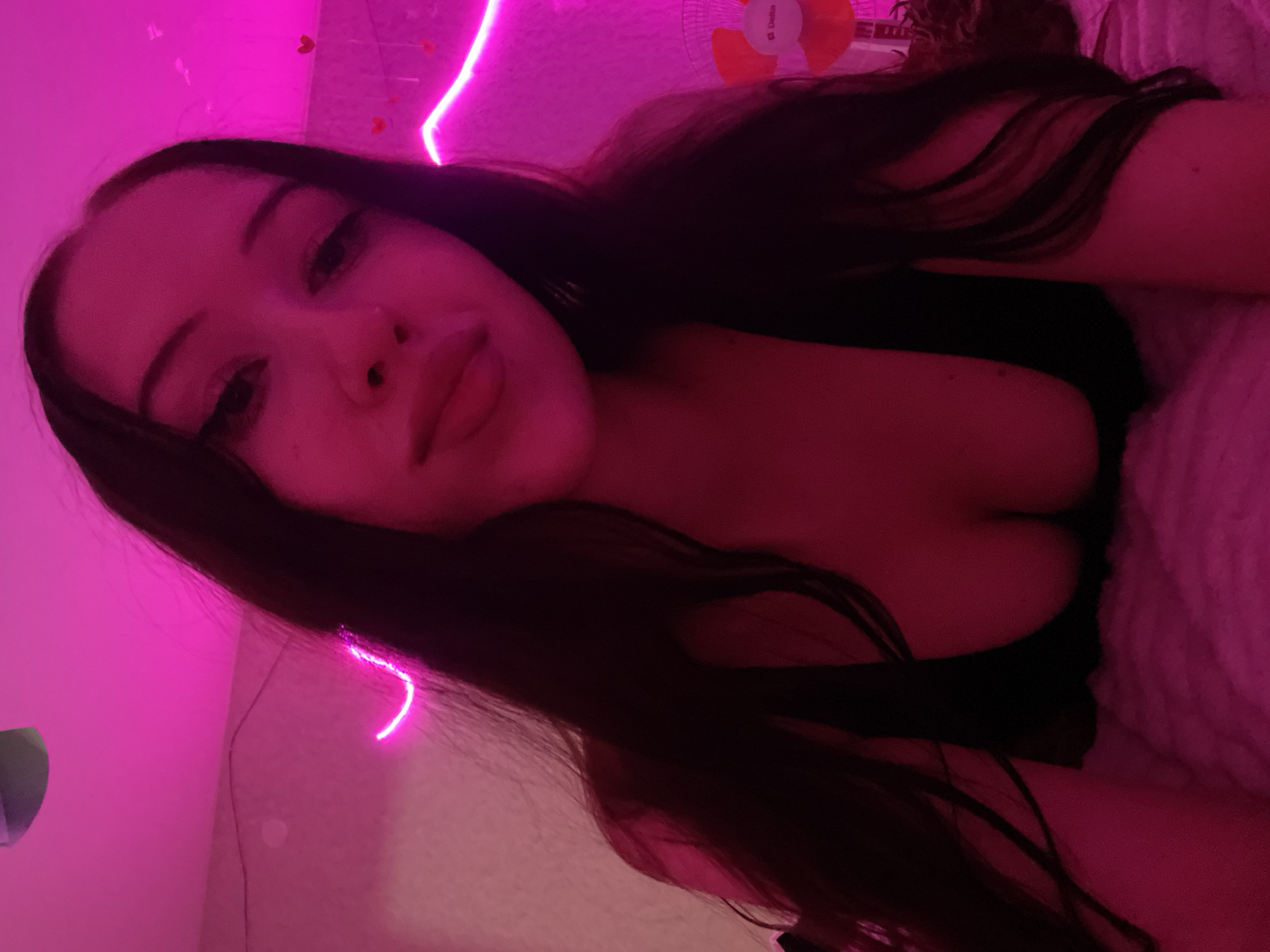 A young woman with long, dark hair is lying down, illuminated by pink neon lights. She is wearing a black top, and her cleavage is visible. The background includes a wall with pink neon lighting and some small heart decorations. There is also a small fan visible on the right side of the image. The setting appears to be an indoor bedroom. The image does not provide any information to determine the location in the city of Votkinsk, Russia.