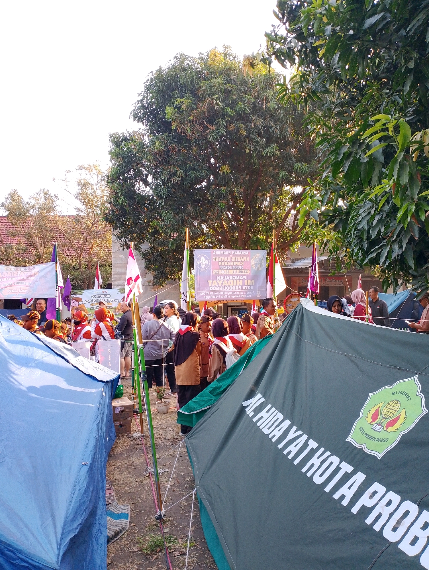 The image shows an outdoor event, likely a gathering or camp, with tents set up. In the foreground, a dark green tent with white lettering prominently displays "AL. HIDAYAT KOTA PROBOLINGGO" and a crest with a globe and an open book. Several people, many wearing scout-like uniforms (brown and orange), are visible around the tents. Some are standing, talking, and observing. In the background, more tents, flags (including red and white Indonesian flags), and trees are present. The sky is bright, suggesting it is daytime, possibly late afternoon due to the warm lighting. The ground appears to be dirt and dry leaves. The overall scene suggests an organized activity in a semi-rural or open area within Probolinggo, Indonesia.