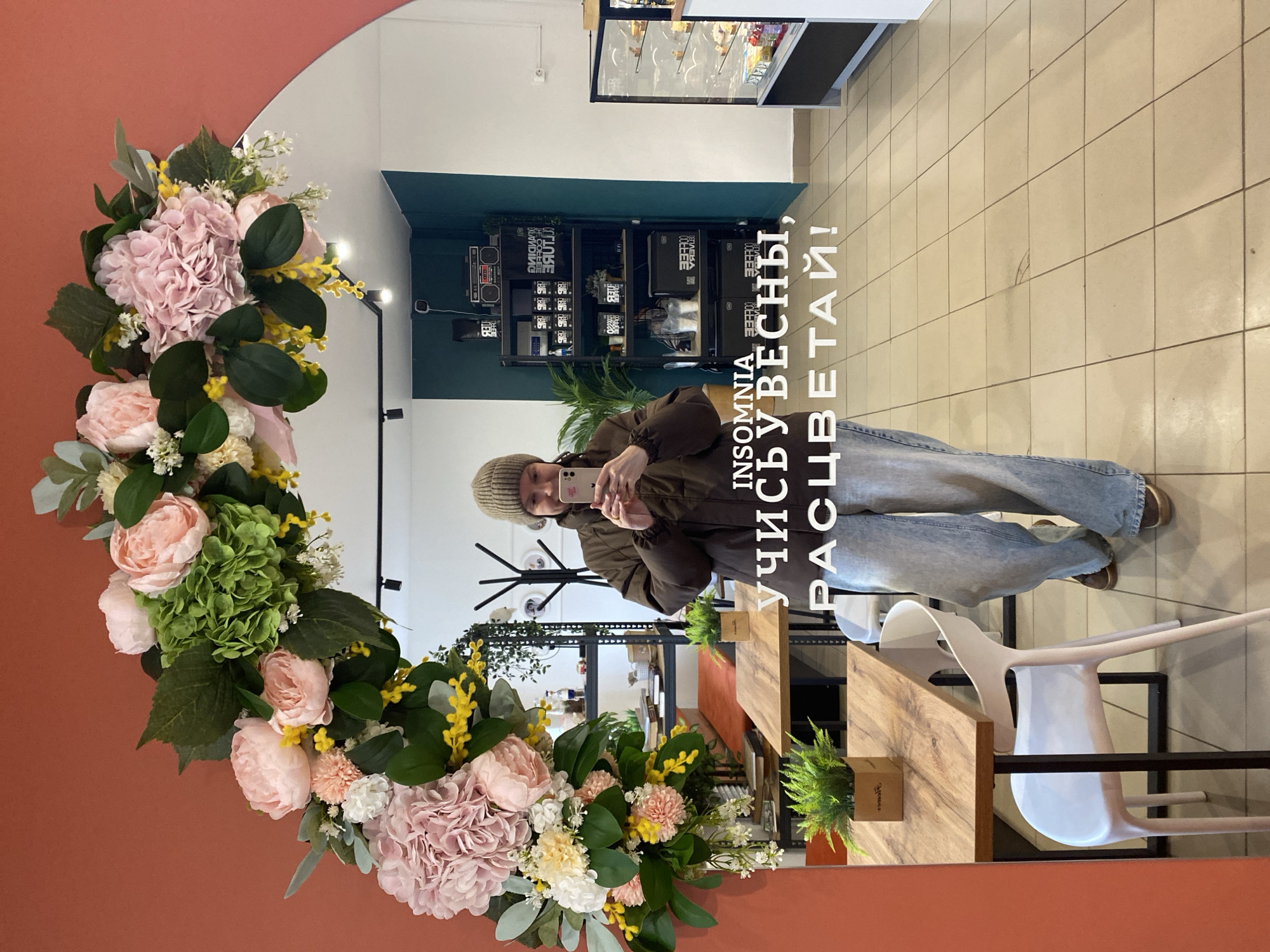 A woman in a brown hat and jacket takes a selfie in a mirror adorned with pink and yellow flowers. The mirror is oval and surrounded by a peach-colored wall. Behind the woman and the mirror, the scene reflects a cafe interior. There are shelves filled with coffee products, and a table with white chairs is visible. The text "INSOMNIA, ЧАЙКОВСКИЙ, РАСЦВЕТАЙ!" is superimposed on the image.