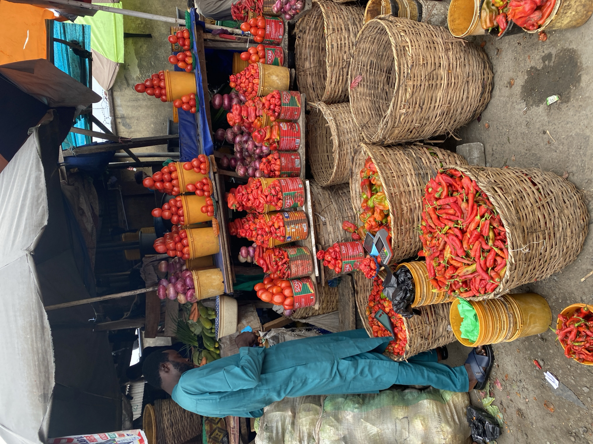 A street-level view in Lekki, Nigeria, shows a man in a teal outfit standing near a vendor's stall. The stall prominently displays tomatoes, red onions, and red peppers, arranged in buckets and stacked for sale. Baskets filled with red peppers are also visible. The environment appears to be a bustling marketplace.
