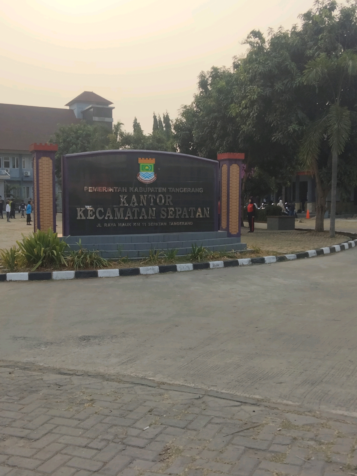 The image shows a large, dark sign in front of a building in Sepatan, Indonesia. The sign has text in Indonesian, reading "PEMERINTAH KABUPATEN TANGERANG KANTOR KECAMATAN SEPATAN" and "JL. RAYA MAUK KM 11 SEPATAN TANGERANG". This translates to "TANGERANG REGENCY GOVERNMENT SEPATAN DISTRICT OFFICE" and "JL. RAYA MAUK KM 11 SEPATAN TANGERANG". The sign is flanked by two decorative pillars. Behind the sign, a building with a grey facade and a dark roof is visible, with trees and other vegetation in the foreground. Several people are seen in the background, some standing near the building and others on motorcycles. The scene appears to be an outdoor administrative building in a semi-urban or suburban area.