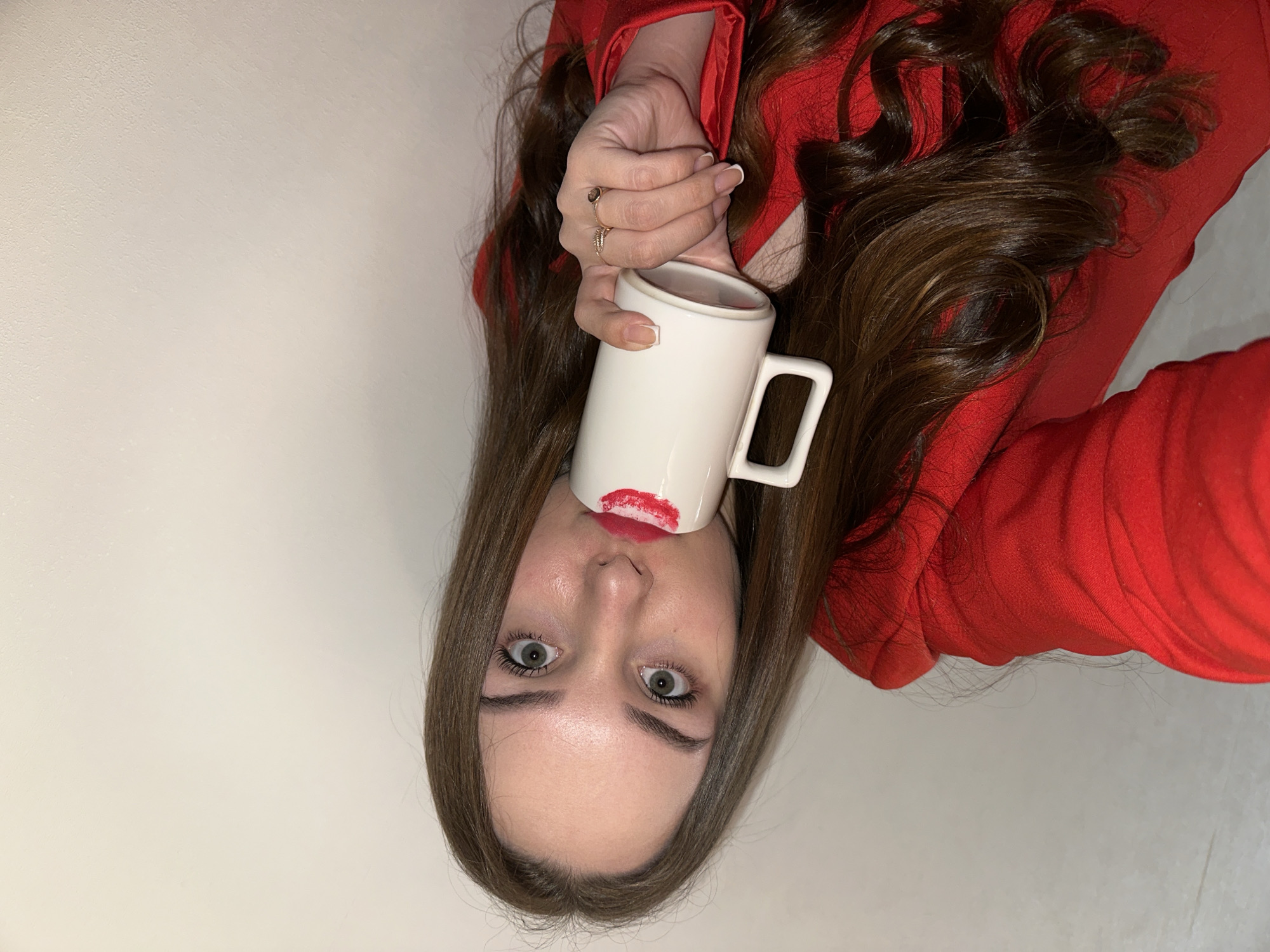 A woman is holding a white mug to her lips, leaving a red lipstick mark on the cup. She has brown hair and light eyes. She is wearing a red top and two rings on her left hand. The background is a light beige wall. The location in Chaikovsky, Russia, cannot be determined from the visual data.