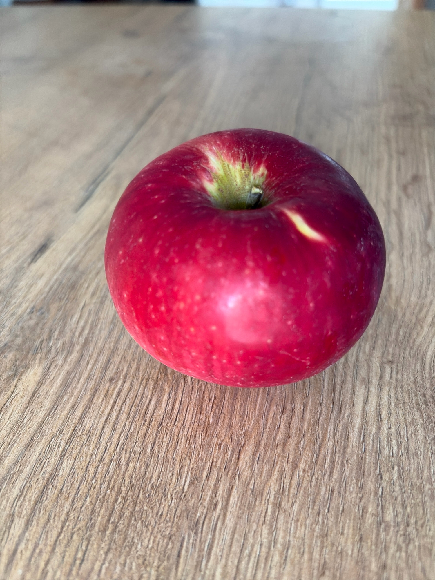 A single red apple rests on a wooden table. The apple is positioned slightly off-center to the right, with its stem area pointing towards the top left of the frame. The table surface displays a prominent wood grain pattern. The lighting appears to be natural, casting soft shadows. No people or other objects are visible in the image. The scene is devoid of any specific urban context, making it impossible to place it within the city of Oxentea, Moldova.