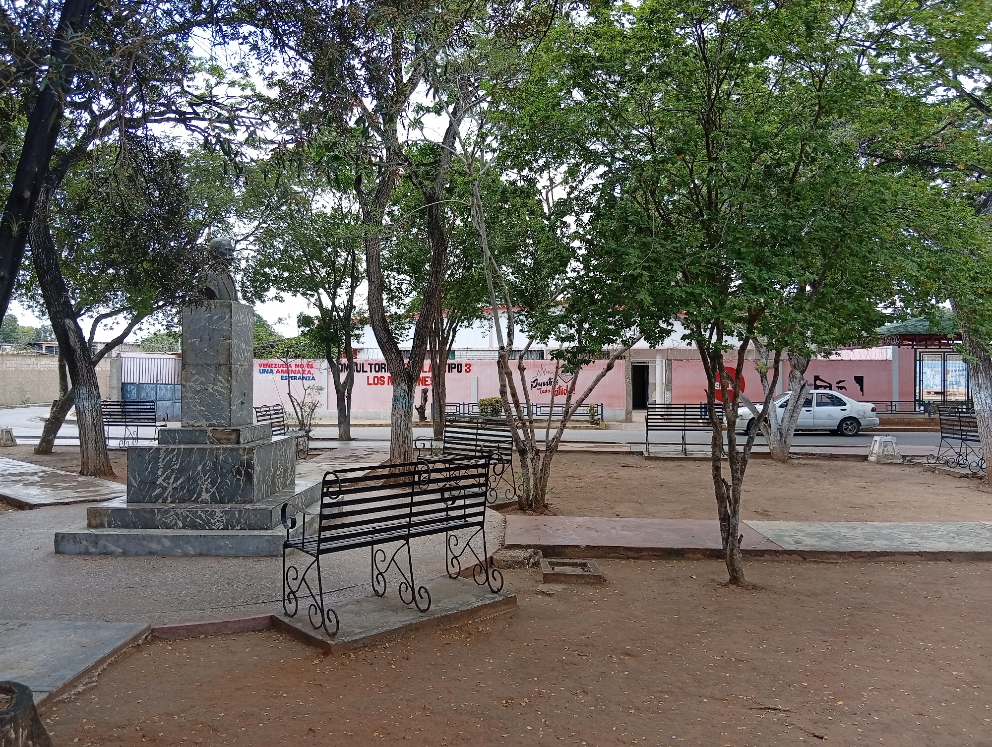 The image depicts a public park in Juan Griego, Venezuela, featuring a monument with a bust at its center. The monument is surrounded by several black metal benches with decorative scrolled legs, some of which are occupied by people. The park is characterized by a dirt ground cover with scattered leaves and some paved pathways. Lush green trees with visible branches and foliage are present throughout the scene, casting shadows on the ground. In the background, a pink building with windows and a green roof is visible, along with a white sedan parked on the street. The sky is partly cloudy.