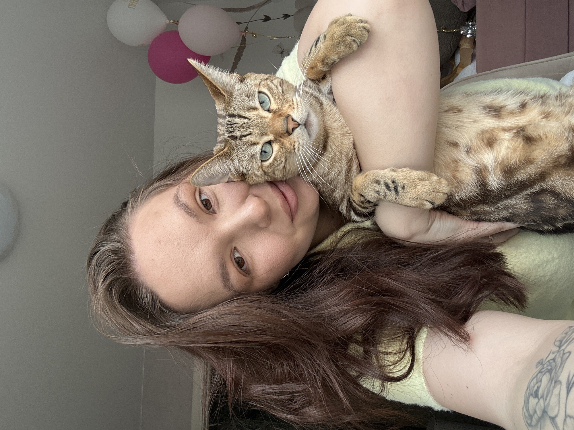 A female subject with long, dark brown hair and light skin is depicted indoors, holding a brown and tan striped tabby cat with blue-green eyes. The woman, wearing a light yellow top, has a floral tattoo on her right upper arm and a subtle smile as she looks towards the camera. The cat is held close to her, resting its front paws on her arm, and stares directly forward. The background consists of a light-colored wall with faint patterning, and in the upper right, three balloons are visible: one white with partial Russian text, one fuchsia, and one light pink. A portion of pink upholstered furniture is present in the lower right. The scene is situated in the city of Chaikovsky, Russia.