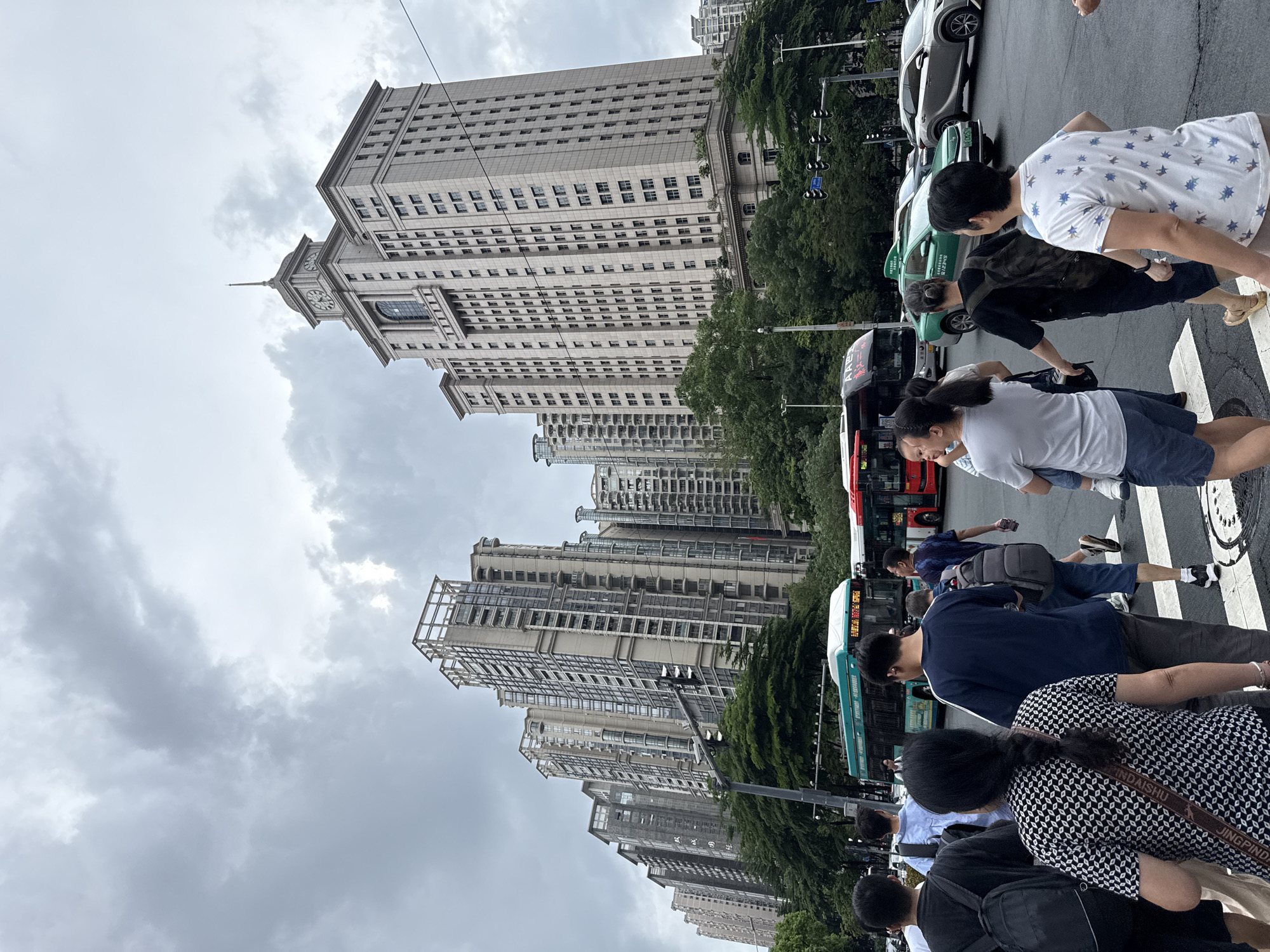 The image depicts a street scene in Tianhe District, China, under an overcast sky. A pedestrian crosswalk is occupied by several individuals walking in various directions. A woman in a black and white patterned top with a crossbody bag is walking away from the camera. To her right, a man in a dark blue t-shirt and grey shorts is also moving away. Behind them, a person in a light blue shirt carries a backpack. In the middle ground, a green bus with the number 101 visible on its side is stopped at the intersection. To the right of the green bus, a red bus is also present. Further right, a green taxi is visible with license plate 粤B CG2515. Several tall, modern apartment buildings with numerous windows dominate the background. The largest building, on the right, is a distinct structure with a clock tower at its peak. Trees line the street between the buildings and the road. Traffic lights are visible at the intersection.