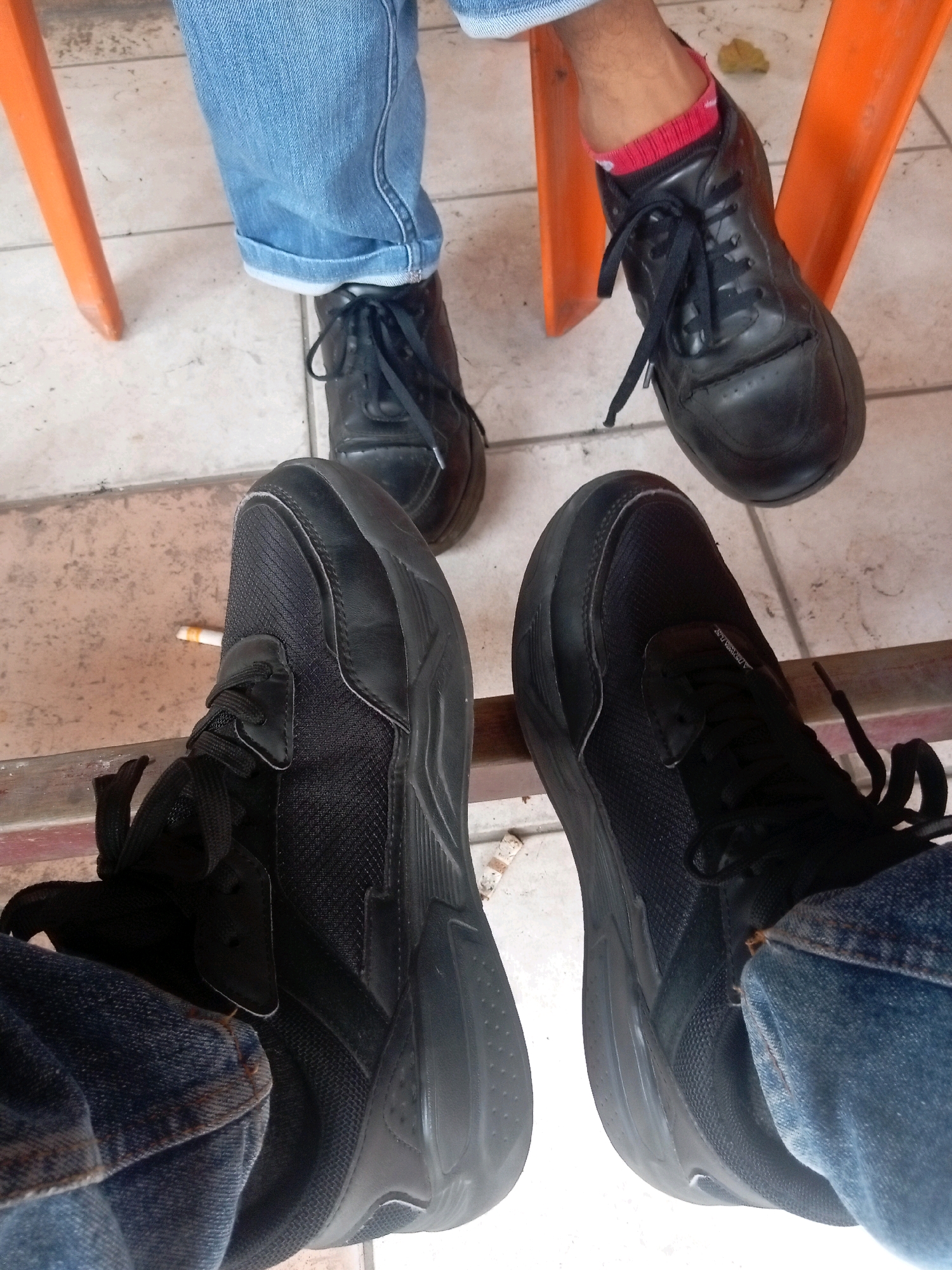 The image shows a close-up view of two people's legs and feet, sitting in what appears to be an outdoor or semi-outdoor setting. One person on the left is wearing blue jeans and black athletic shoes. The other person on the right is also wearing blue jeans and black shoes, with red socks visible on their right foot. Both individuals appear to be wearing casual attire.
The environment includes an orange chair on the left side of the frame, suggesting they are seated. The ground is tiled. A small, lit cigarette butt is visible on the floor near the athletic shoe on the left. The overall impression is of a casual, everyday moment, possibly at a cafe, waiting area, or public space.
Given the request for location in Batam, Indonesia, the tiled flooring and the type of chairs could be consistent with some public spaces or eateries in the city. However, without further visual cues or context, it is not possible to definitively pinpoint the exact location in Batam.