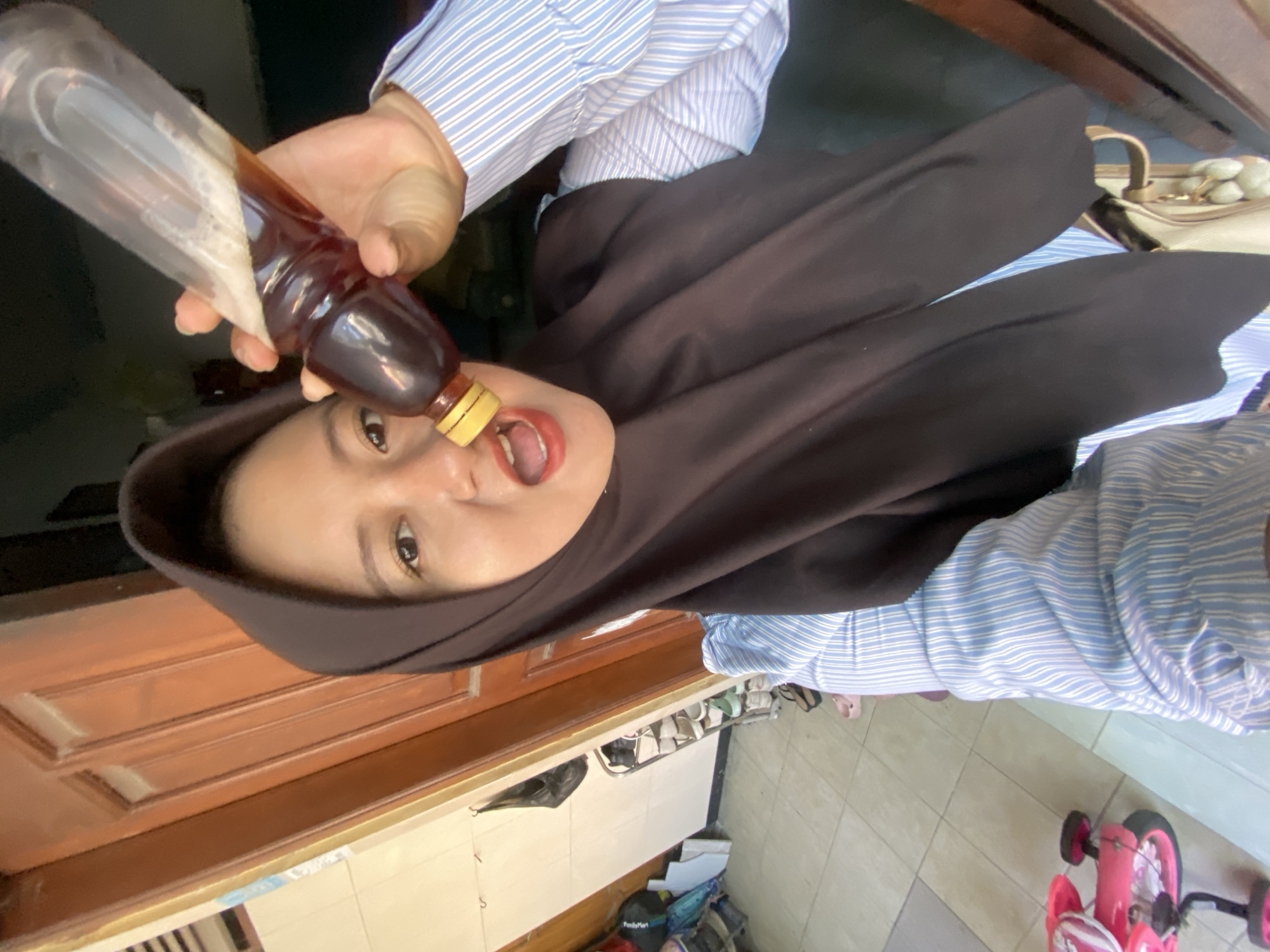 The image shows a woman in a black hijab and a blue and white striped shirt holding a clear bottle with a dark liquid, possibly honey or soy sauce, and tilting it towards her open mouth. She appears to be tasting or drinking from the bottle. Her eyes are wide open, and her mouth is slightly ajar. The scene appears to be outdoors, with a wooden door in the background and tiled flooring. There are shoes and bags on the floor, and a pink toy scooter is visible in the lower right corner, suggesting a residential area. The lighting suggests it might be daytime. The woman's expression is neutral or slightly amused. No text is clearly visible in the image.