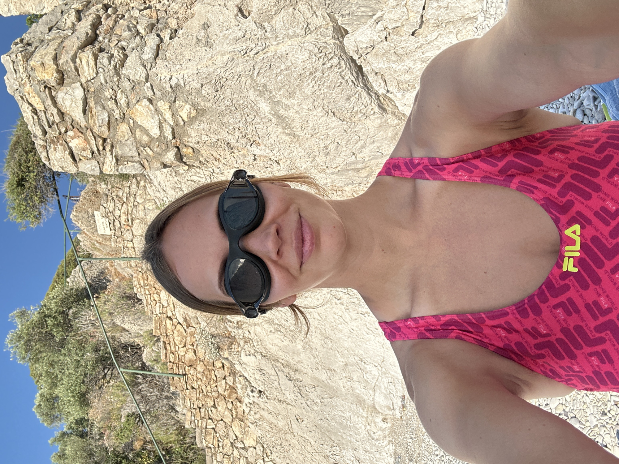 A young woman is captured in a sunlit selfie, likely taken in a coastal area of Fethiye, Turkey. She is positioned at an angle, looking directly at the camera with a gentle, relaxed smile. Dark swimming goggles are worn on her head. She is clad in a bright pink one-piece swimsuit featuring a repeating pattern of the brand name "FILA" and the phrase "ONE WORLD," with an additional yellow "FILA" logo prominently displayed near her right shoulder strap.
The background consists of rugged, light-colored rocky cliffs and sparse green vegetation under a clear blue sky, suggesting a bright and warm day. Some green metal rods or supports are also visible further back in the landscape. The ground immediately behind her appears to be a mix of rocks and pebbles, typical of a rocky beach or shoreline. The scene conveys a sense of leisure and enjoyment of the natural, sunny environment.