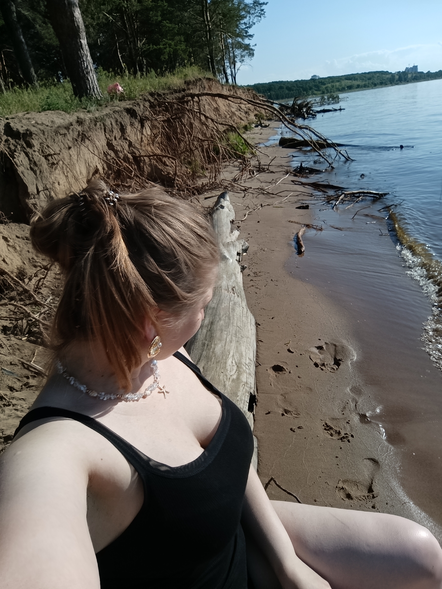 A young Caucasian woman with light skin and auburn hair is seated on a sandy beach, facing away from the camera and looking towards a body of water. She is wearing a black tank top and a pearl necklace with a starfish pendant. Her hair is pulled up in a messy bun, adorned with a sparkly hair clip. The background shows a sandy shore with footprints, scattered driftwood, and a steep, eroded embankment covered in grass and tree roots. Beyond the embankment, a dense forest of green trees stretches into the distance. The water is calm and reflects the clear blue sky. A few buildings are visible on the far horizon, along with trees. The overall scene suggests a sunny day at a natural beach.