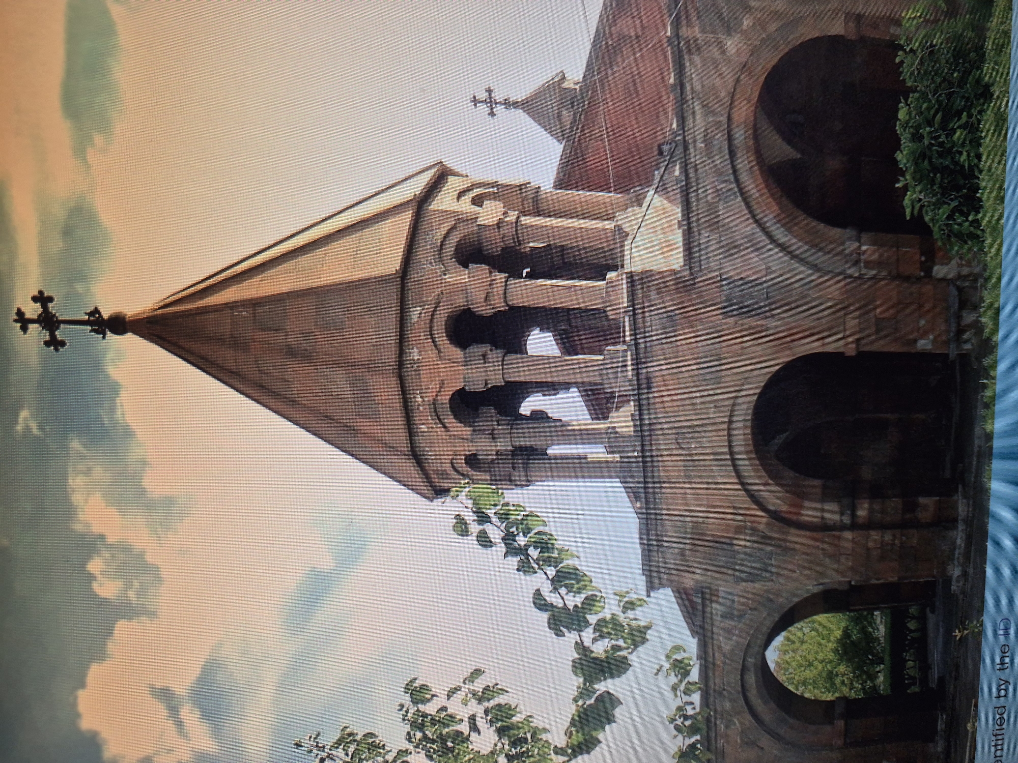 The image depicts the Church of Saint Hripsime in Yerevan, Armenia. The photo is taken from a low angle, showcasing the church's bell tower and its distinctive conical roof, topped with a cross. The building is made of stone and has an aged appearance. The bell tower features a series of arches. The background includes a light blue sky with light clouds. The bottom of the image features a small tree branch with leaves in the foreground. The building has a series of arches.