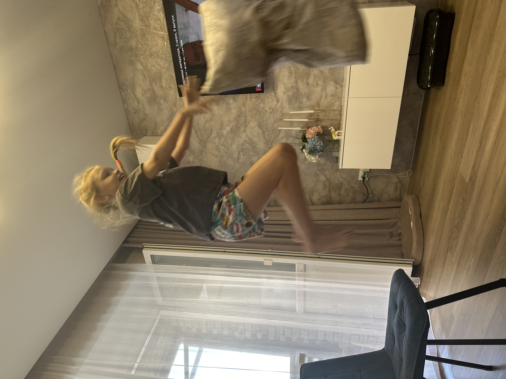 The image shows a young person in mid-air, appearing to be jumping or falling, with a pillow overhead. The person is wearing a grey t-shirt and colorful shorts. They have blonde hair and appear to be smiling. The scene is indoors, with a television screen displaying text that reads "Сокровища императора, 3 сезон, 6 выпуск, канал ТНТ". To the right of the television is a white cabinet with decorative candles and flowers. The room has a textured wallpaper and a wooden floor. A window with sheer curtains is visible in the background. A dark chair with metal legs is in the foreground. The image is oriented vertically, suggesting it was taken with a phone in portrait mode. The lighting suggests it is daytime. The overall impression is of a playful or energetic moment captured indoors.