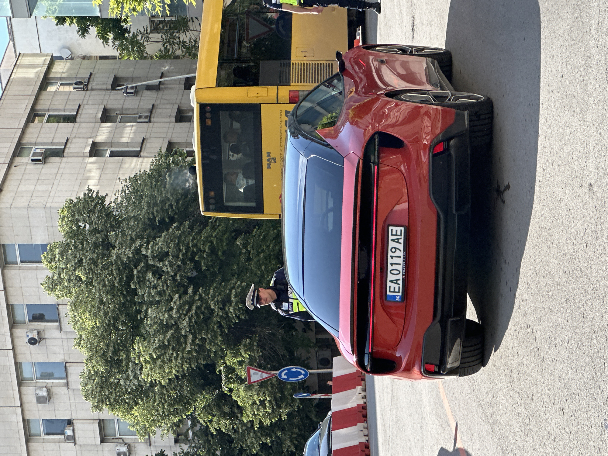 A red Polestar 3 is stopped on a street in Veliko Tarnovo, Bulgaria, next to a yellow bus. The license plate of the Polestar 3 is EA0119AE. A Bulgarian police officer wearing a white hat and uniform is standing next to the car. Traffic signs are visible next to the car. In the background is a light-colored building and a tree. The day appears to be sunny.