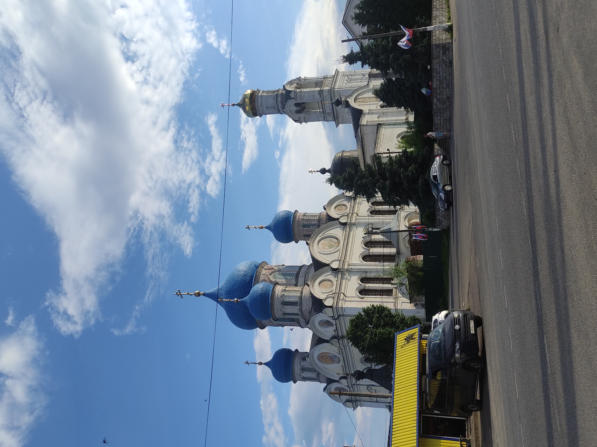 The image shows a large white church with multiple blue domes and a single gold dome on a bell tower, located in the city of Rogachovo, Russia. The church architecture features intricate detailing, arched windows, and crosses on its domes. In the foreground, a black van is parked in front of a yellow building with a corrugated metal roof. Across the street, a silver sedan is parked and a person is standing near a low brick wall decorated with a wreath. Several evergreen trees are scattered around the church and the surrounding area. The sky is blue with scattered white clouds, and a few birds are visible in the sky. Power lines stretch across the upper portion of the image. The road in front of the church and the yellow building appears to be asphalt. The scene depicts an exterior view of a religious site and its immediate surroundings on a clear day.