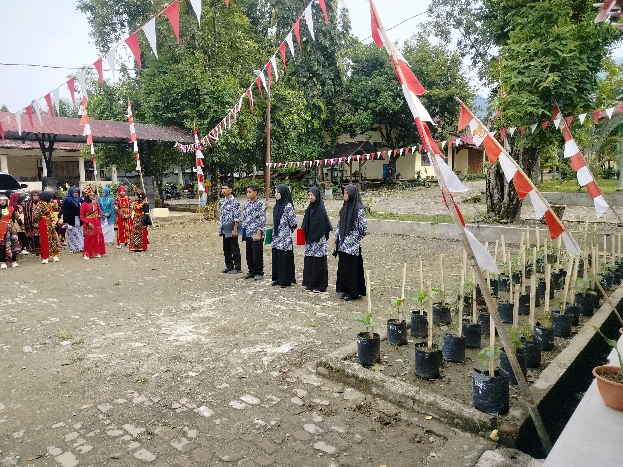 The image shows an outdoor scene in Purwodadi, Indonesia, possibly a school or community event. In the foreground, a group of people are lined up. On the left, a group of individuals in traditional colorful attire are gathered, some wearing head coverings. To their right, four young people, two males and two females wearing headscarves, stand in a line, looking forward. All four are wearing matching printed shirts and dark pants or skirts. Behind them, a few other individuals are visible, some also in traditional clothing. Red and white triangular flags are strung across the scene, along with larger red and white flags on bamboo poles, suggesting a national celebration or event. The ground is a mix of dirt and brick paving. In the background, there are buildings with red-tiled roofs, trees, and more red and white flag decorations. To the right of the lined-up individuals, a row of small plants in black plastic bags, each supported by bamboo stakes, is arranged along a concrete edge. The sky is overcast.