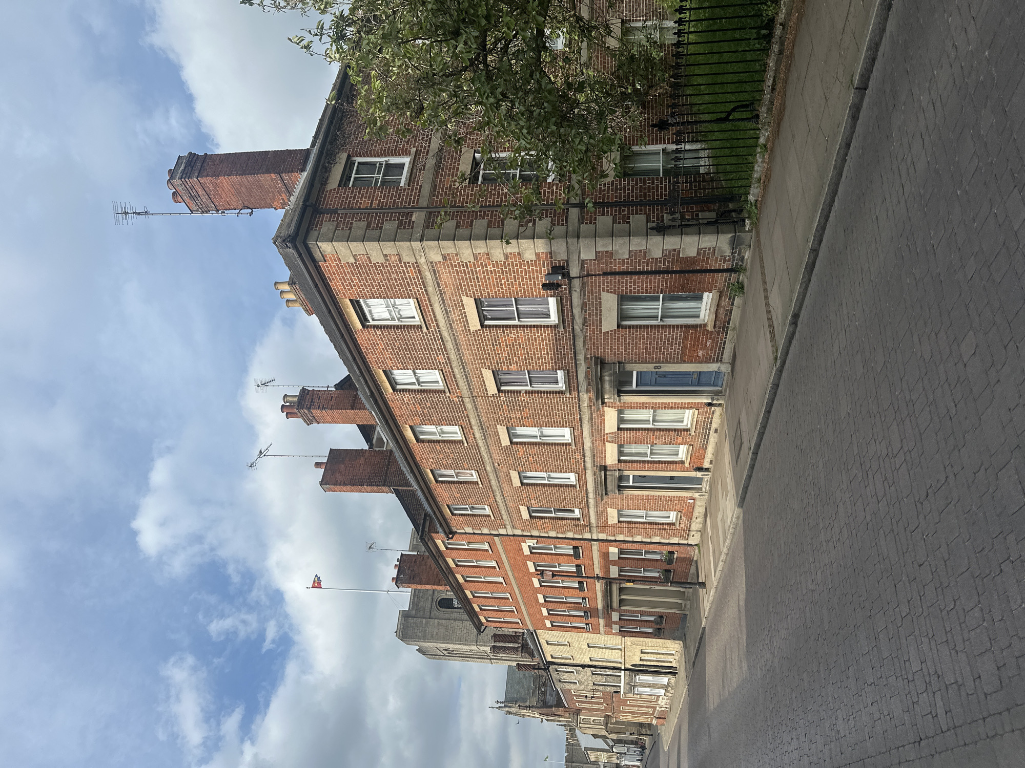 The image depicts a street in West Suffolk, United Kingdom, characterized by brick buildings of varying heights. These buildings exhibit white-framed windows, dark rooftops, and multiple chimneys extending upwards. The building on the left houses an antenna. A flag flies atop a building further down the street. The sky is partly cloudy, with hints of blue visible between the white cloud formations. The street appears to be paved with gray cobblestones. A narrow sidewalk runs alongside the buildings, with a dark wrought-iron fence separating the street from the buildings. A tree is partially visible in the upper right corner. The image is taken from street level. No people are directly visible.