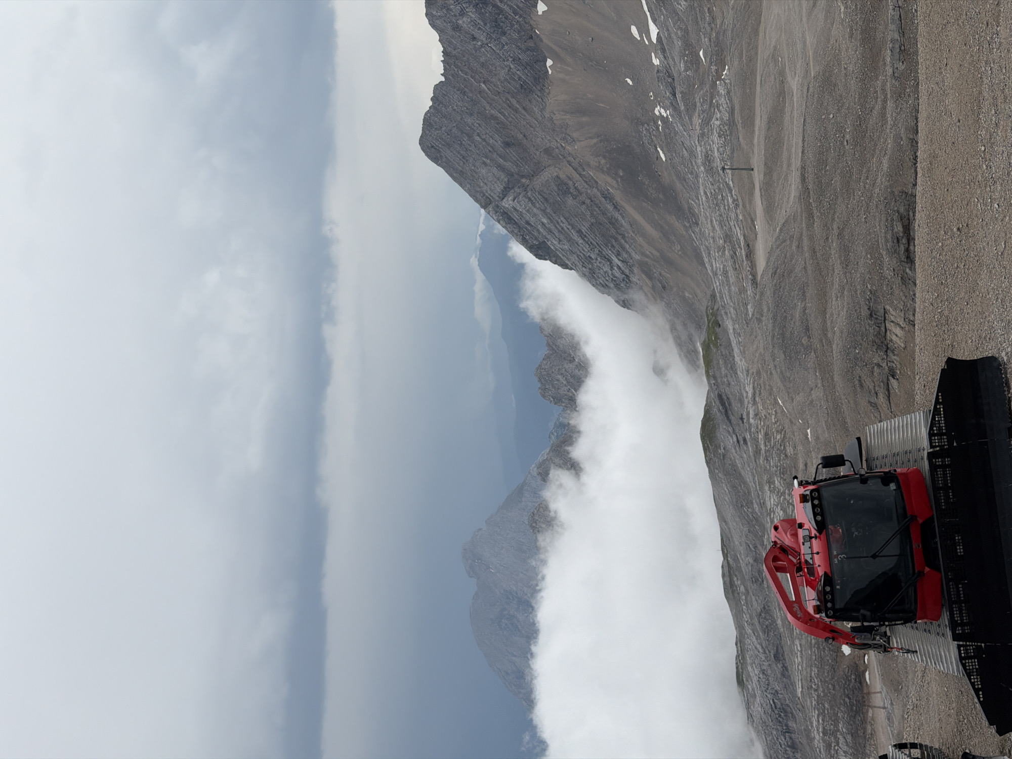 This image depicts a mountainous landscape with a red snowcat vehicle in the foreground. The snowcat, a PistenBully model, is positioned on a gravelly surface, facing towards the viewer. It has a black track system and a red cabin with black-tinted windows. A red arm is extended from the top of the vehicle.
In the background, rugged mountain peaks rise, partially obscured by dense, white clouds that create a dramatic contrast with the grey sky. The terrain is rocky and barren, with some patches of snow visible on the slopes. A solitary pole, possibly part of a ski lift infrastructure, stands in the middle distance. The overall atmosphere is overcast and moody.
The location is the Zugspitze mountain, the highest peak in Germany, situated near Garmisch-Partenkirchen. This specific viewpoint is likely from a higher elevation area of the mountain, possibly a ski run or a viewing platform, given the presence of the snowcat. The scene captures the raw beauty of the alpine environment under a cloudy sky.