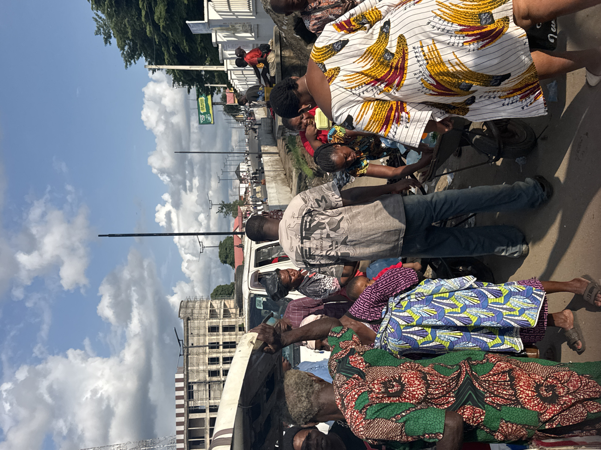 The image captures a busy street scene in Owerri, Nigeria, likely at a market or roadside stall. In the foreground, several people are engaged in various activities. One individual, wearing a grey t-shirt with a graphic print and blue jeans, is seen from behind, looking towards a group of people. A woman in a vibrantly patterned green and red dress stands to the left, facing away from the camera. Next to her, another person, partially visible, wears a blue and purple patterned fabric wrapped around their waist. In the center, a man in a patterned shirt is bent over, possibly tending to goods or assisting someone. A woman in a red top is seated and eating from a yellow container. To the right, a woman in a white dress with a bold yellow and red abstract pattern is captured from the waist down. Behind them, a white van is parked, and construction is visible in the background on an unfinished building. The environment appears to be a street with a sidewalk and road, with streetlights and some trees visible. The sky is blue with scattered clouds. The overall impression is one of daily life and commerce in a Nigerian urban setting.