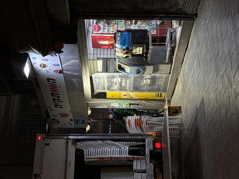 A nighttime street scene features a vending truck parked alongside a sidewalk. The truck is filled with stacked white chairs and orange buckets. A person in a green jacket is visible. Behind the truck, a store or stall displays Coca-Cola products, clear plastic containers, and a yellow Schweppes advertisement. The storefront has a sign with a bright light. The image is rotated 90 degrees counter-clockwise.